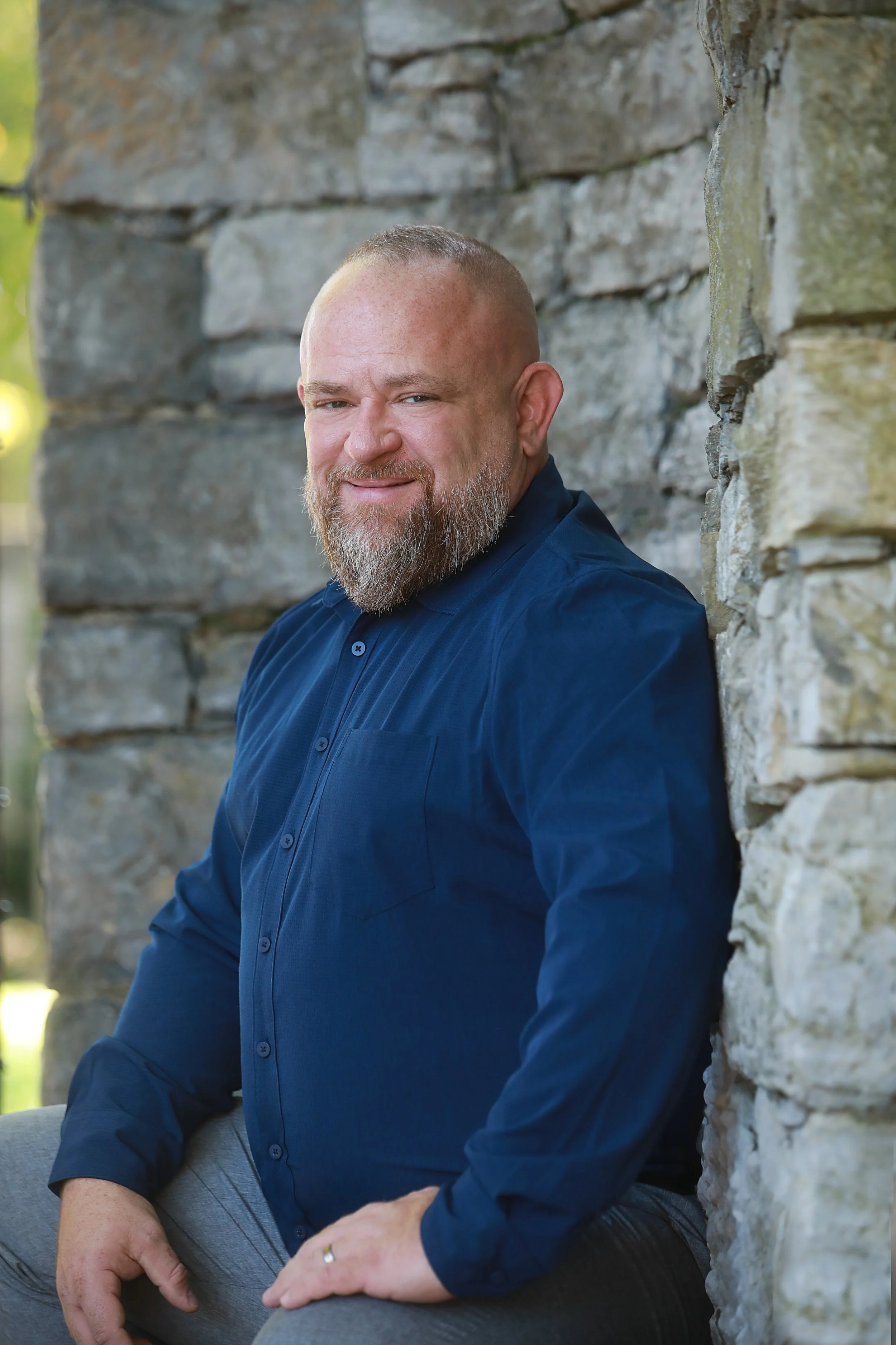 A man with a beard and a shaved head, wearing a navy blue shirt, leaning against a stone wall outdoors.