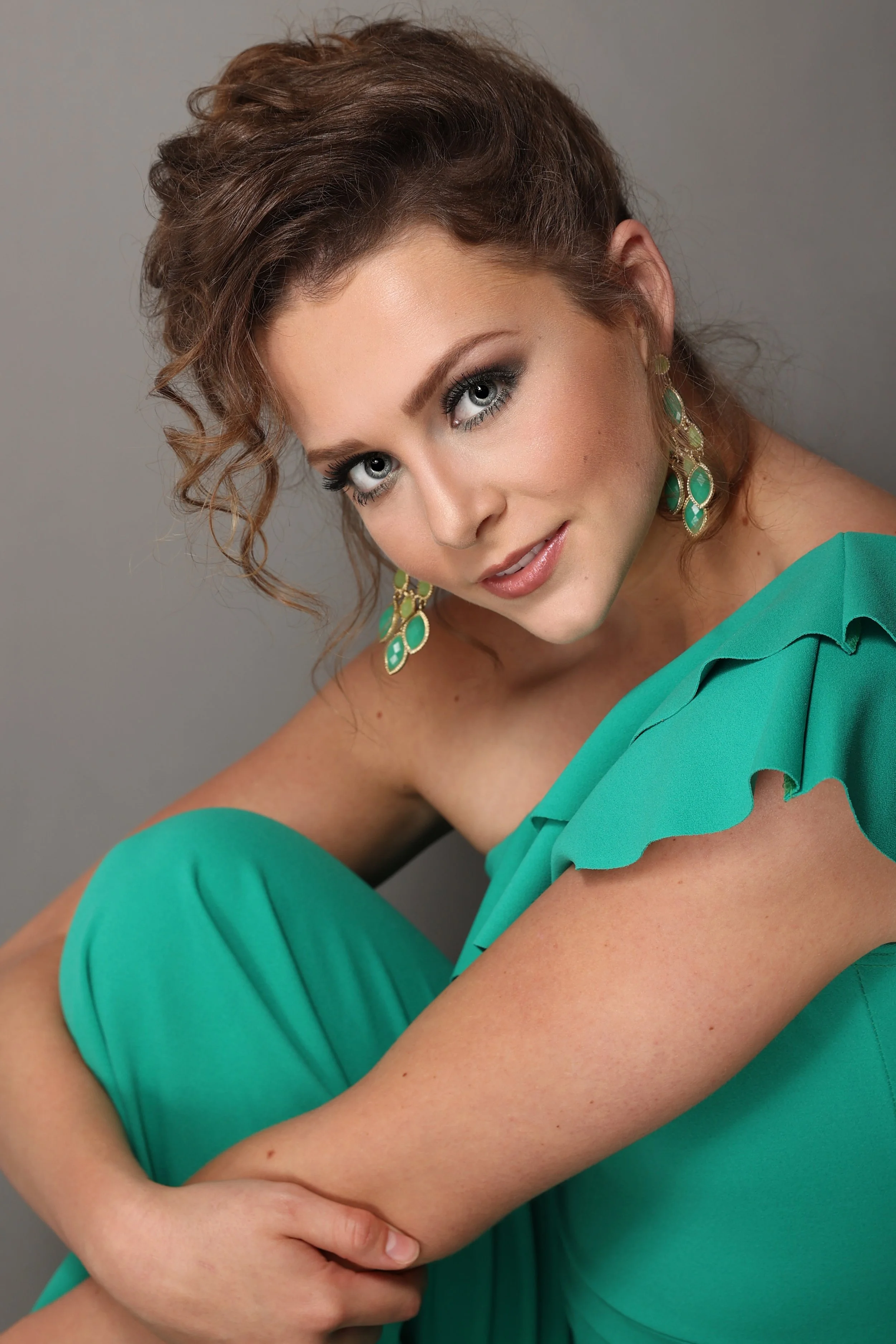 A woman with brown, curly hair and blue eyes, wearing a teal dress with ruffled sleeves and matching dangling earrings, poses against a gray background.