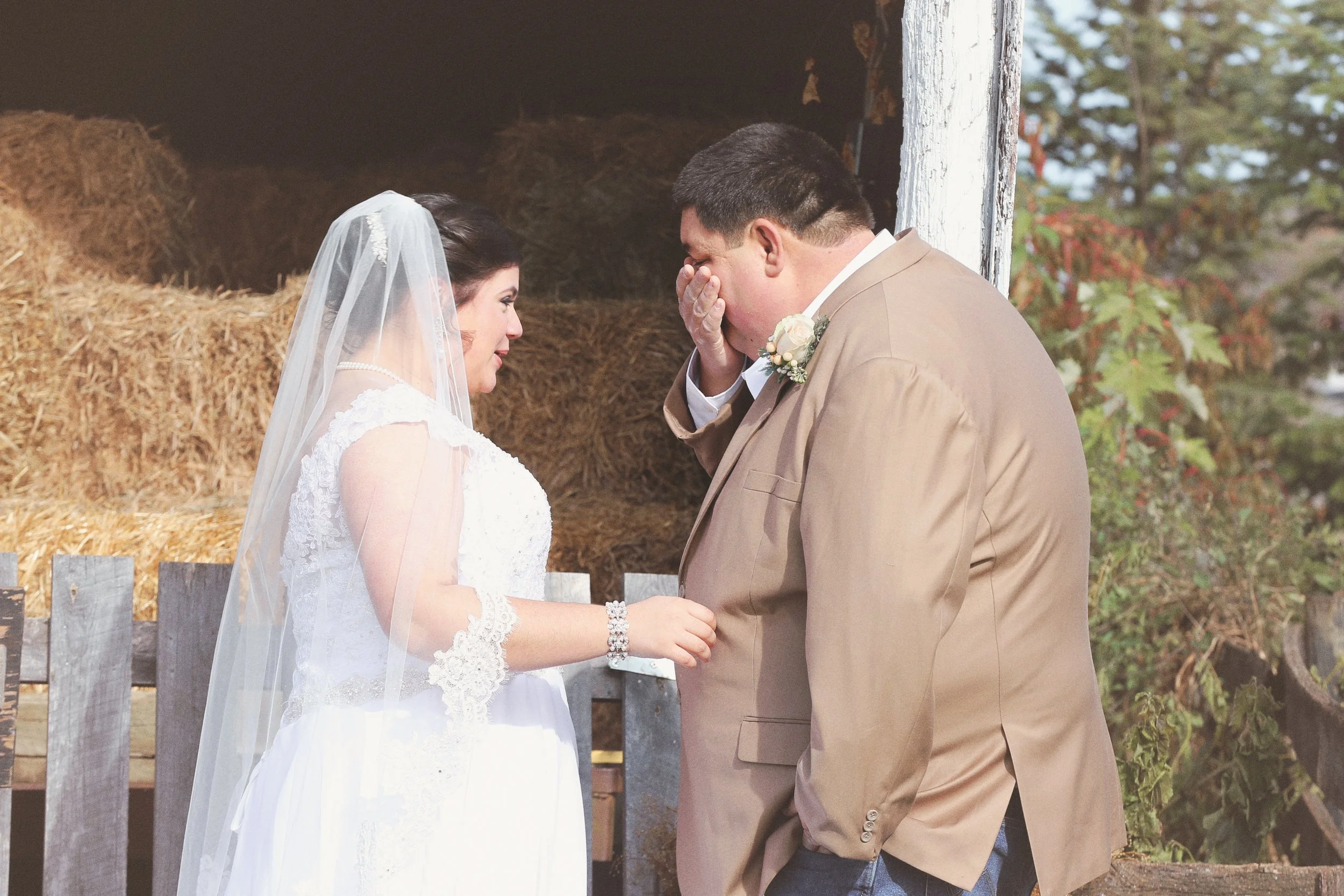 A bride and groom sharing an emotional moment, with the bride smiling and the groom wiping away tears, in front of a barn with hay bales and trees.