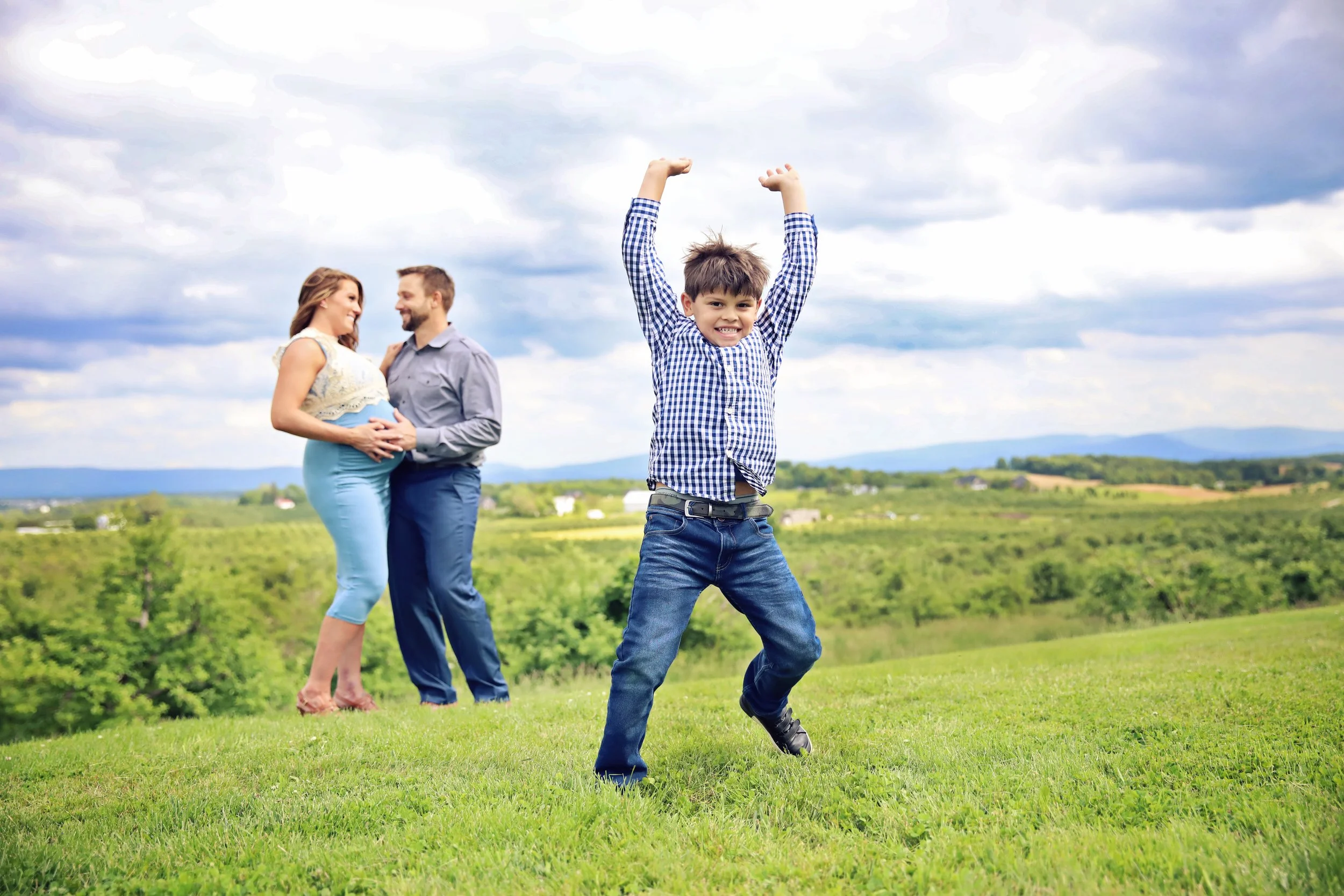 A boy in a checkered shirt and jeans is joyfully jumping for joy in a green field, with a family of three in the background. The family includes a pregnant woman and a man, both smiling and holding each other.