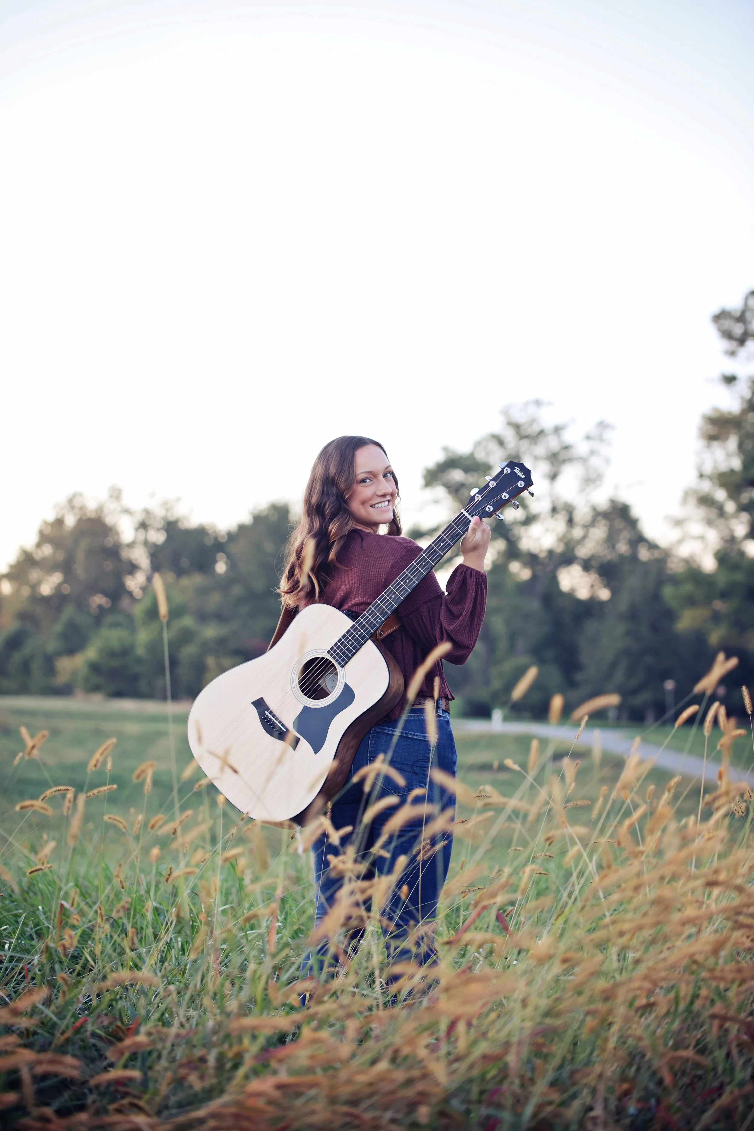 A woman standing in a field with tall grass, holding an acoustic guitar over her shoulder and smiling at the camera, with trees and a clear sky in the background.