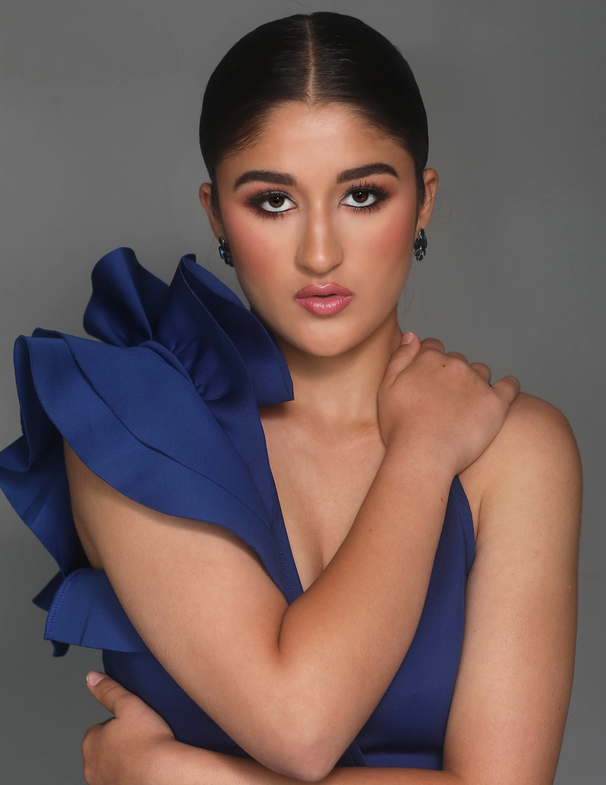 A young woman with dark hair in a sleek style, wearing makeup and a blue dress with large ruffle details on one shoulder, posing against a gray background.