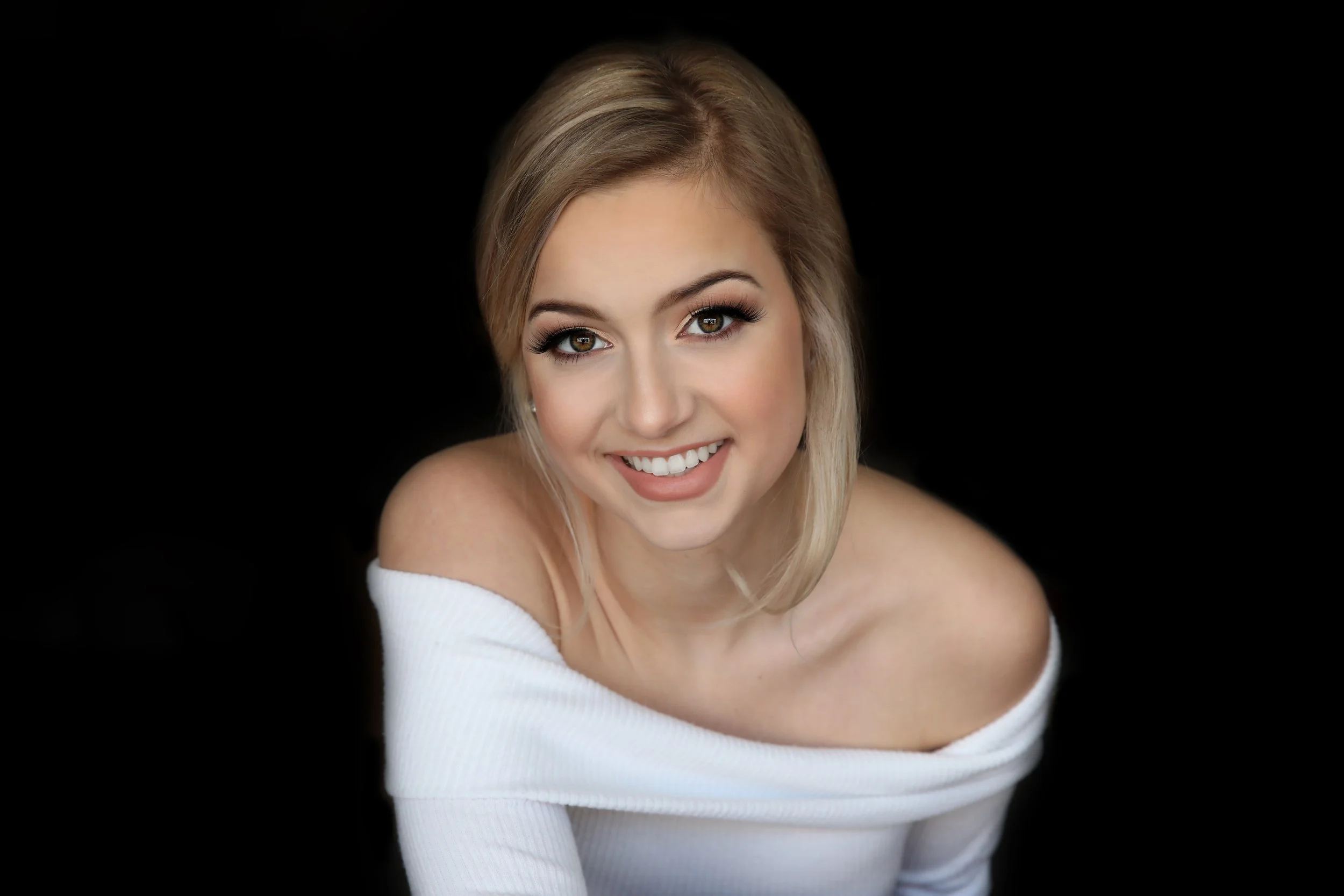 Close-up portrait of a young woman with blonde hair, hazel eyes, and a radiant smile, wearing an off-the-shoulder white sweater against a black background.