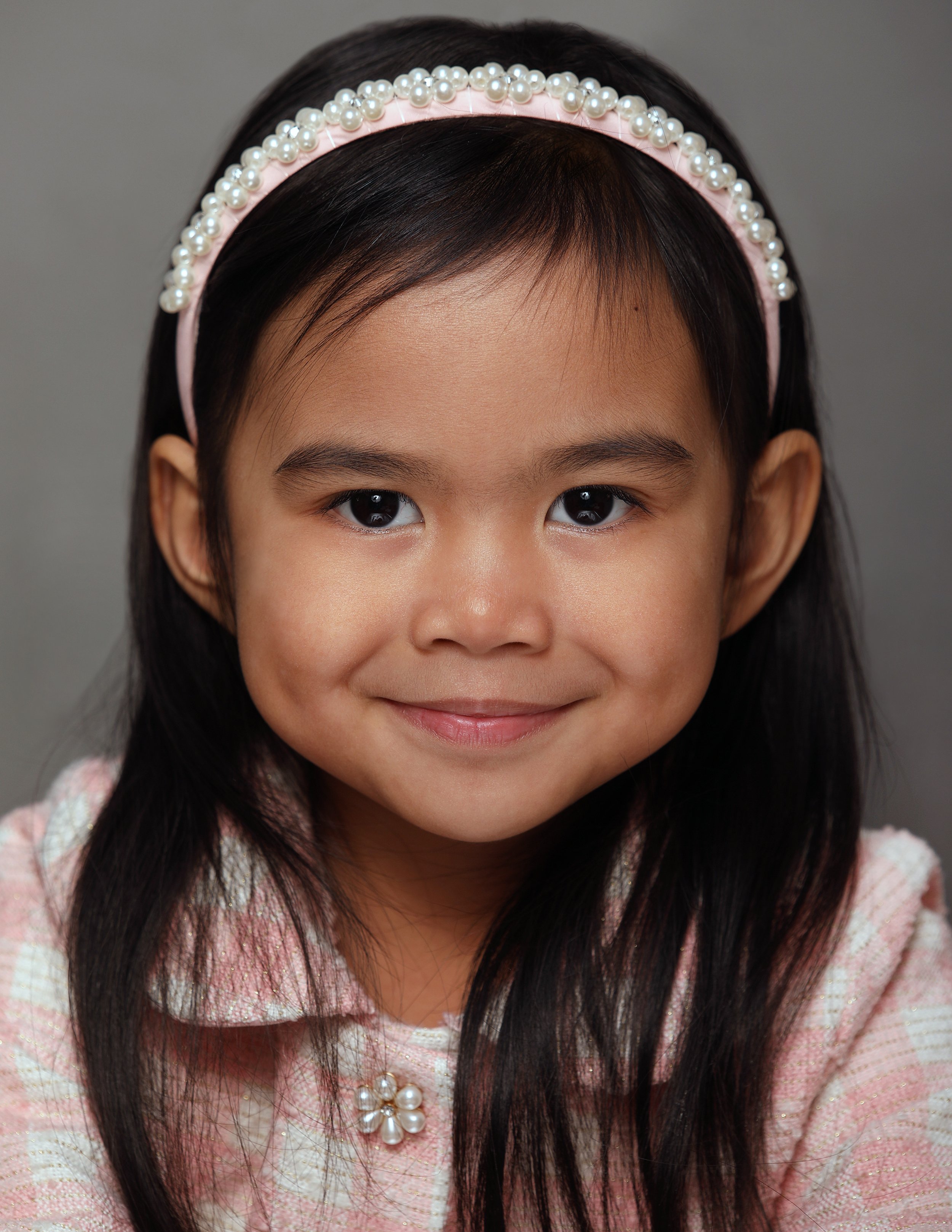 A young girl with long dark hair, wearing a pink headband with pearl embellishments and a matching pink blouse with pearl jewelry, smiling softly.