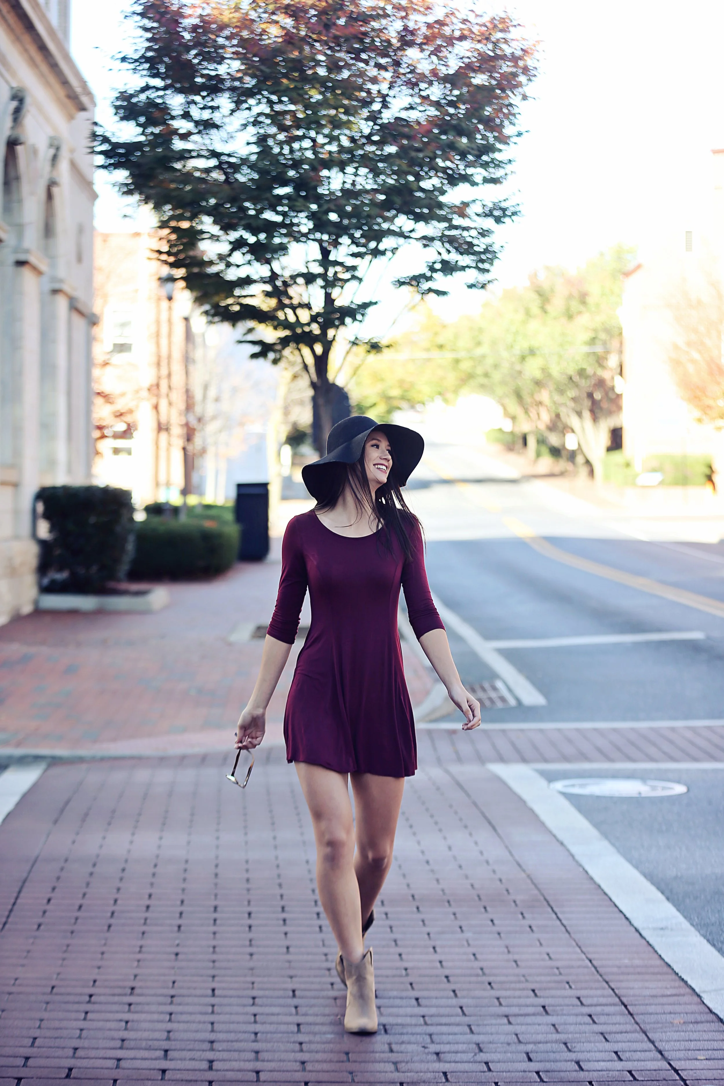 A woman in a burgundy dress and beige high heels walking on a city sidewalk, wearing a large black hat and holding sunglasses, smiling, with trees and a building in the background.