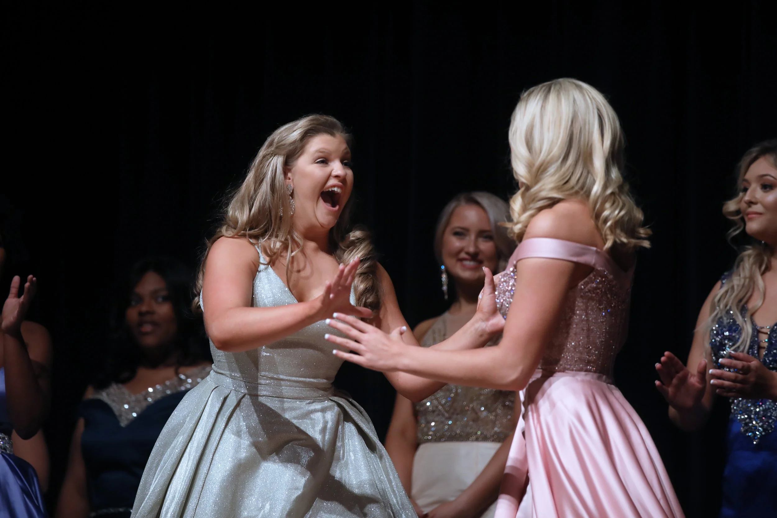 Two young women in evening gowns share an emotional moment, touching hands and smiling, with other women in formal attire in the background.
