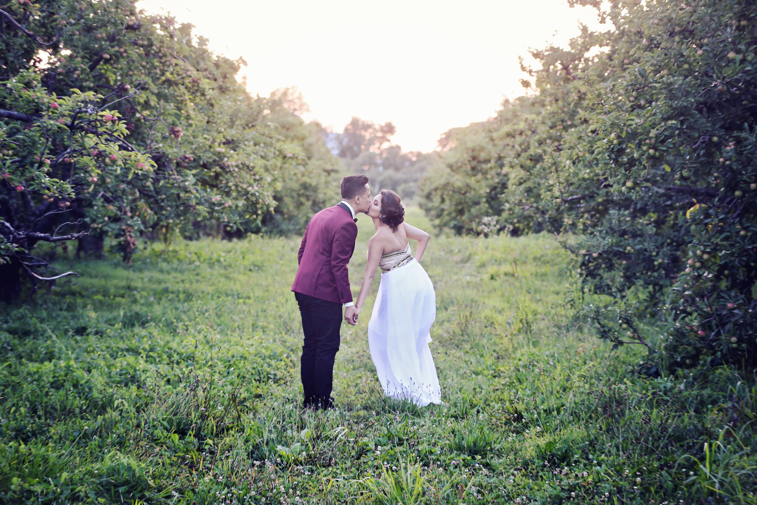 A couple sharing a kiss in a lush green orchard at sunset, holding hands.
