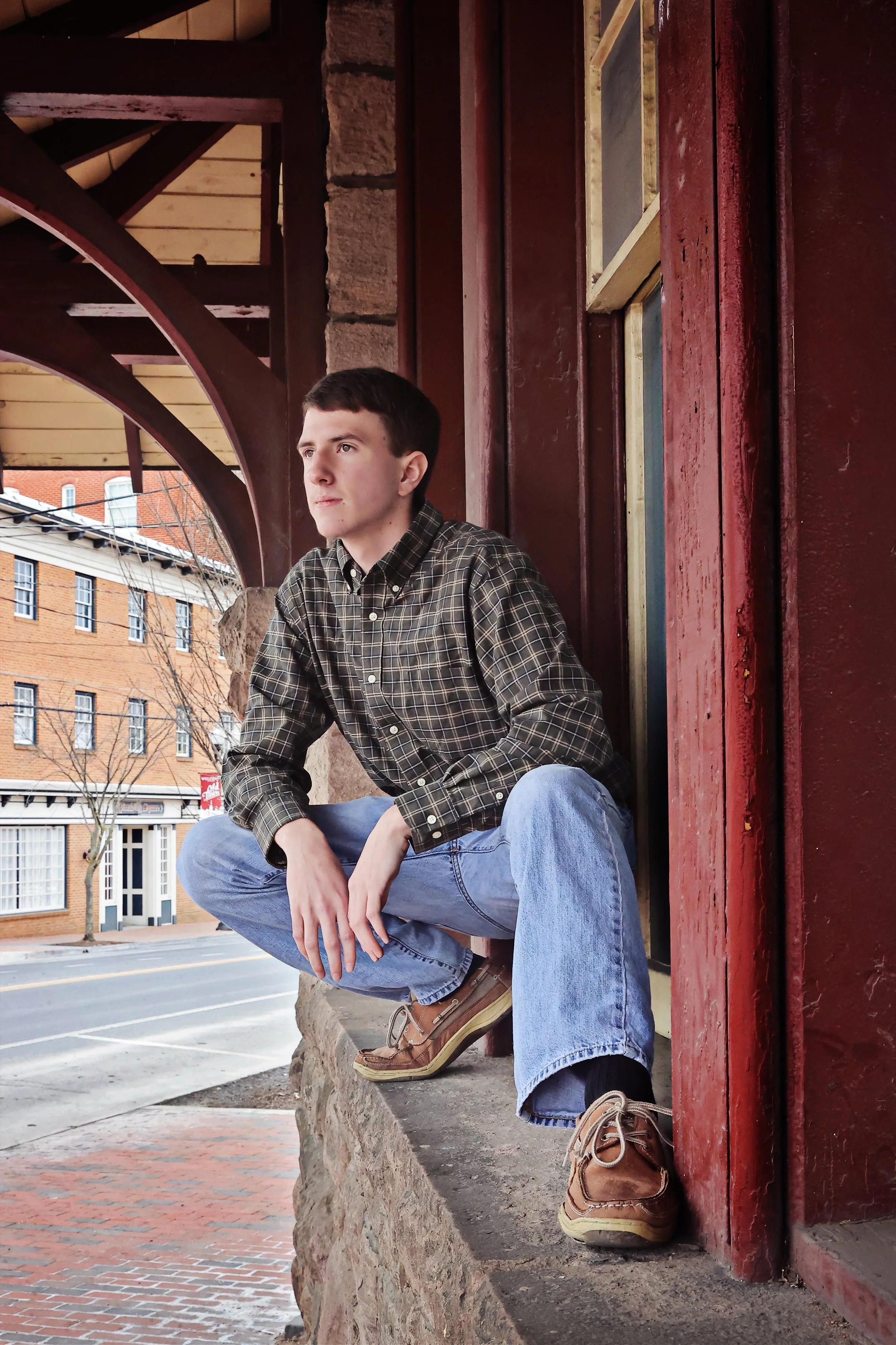 A young man in a plaid shirt and jeans sitting on a stone ledge outside a red brick building with large windows and wooden accents.