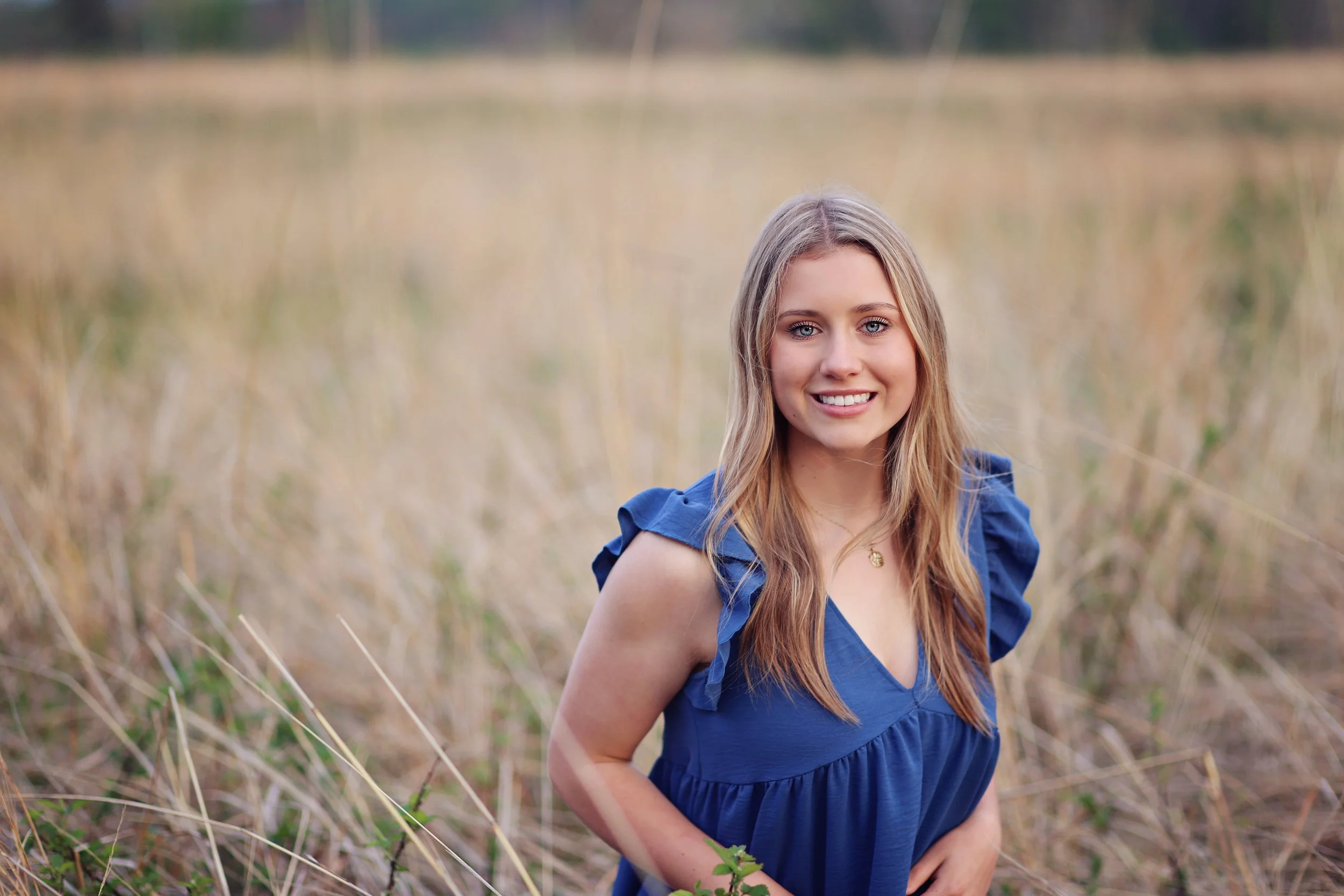 A young woman with long blonde hair smiling outdoors in a field of tall, dry grass, wearing a blue dress.