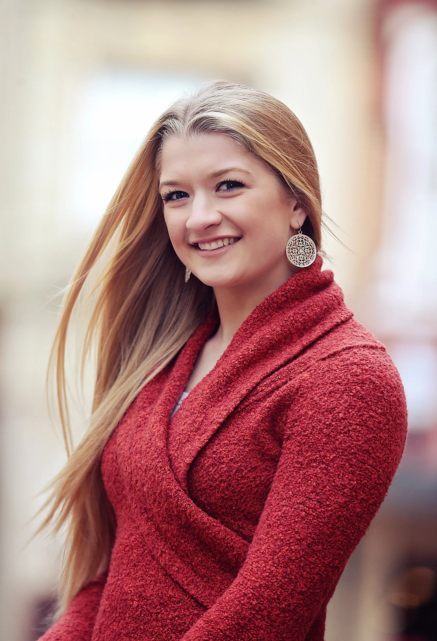 A young woman with long blonde hair, smiling, wearing a red textured coat and large dangling earrings, standing indoors with a blurred background.