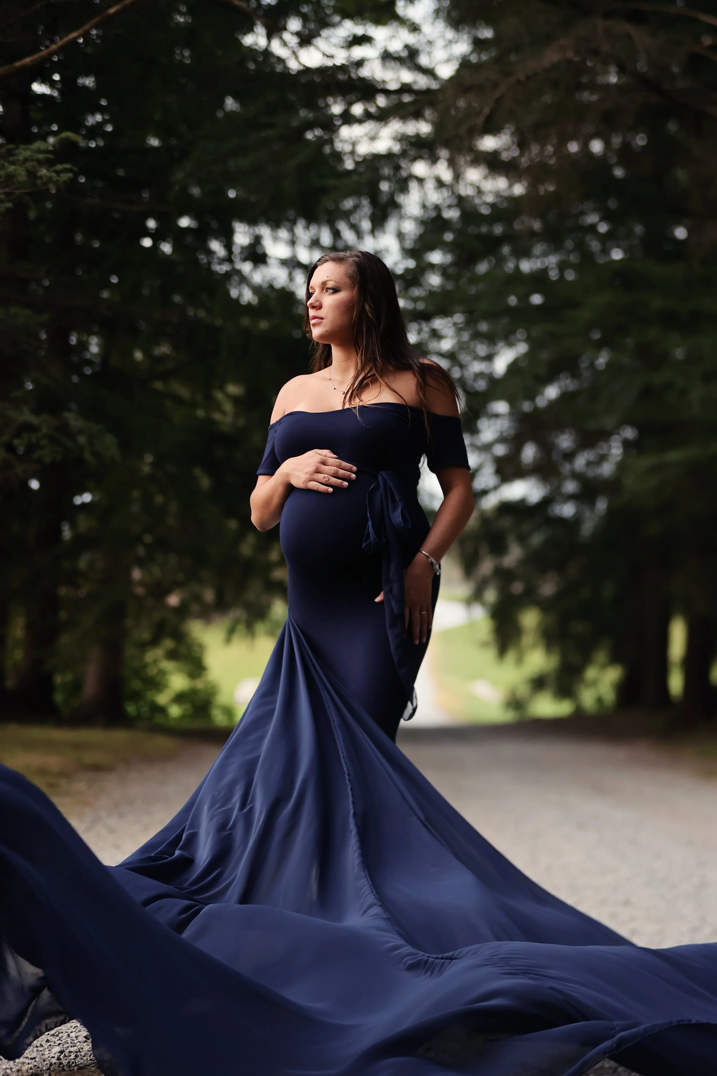 A pregnant woman in a dark blue off-the-shoulder gown standing outdoors on a gravel path surrounded by large trees, holding her belly with one hand.