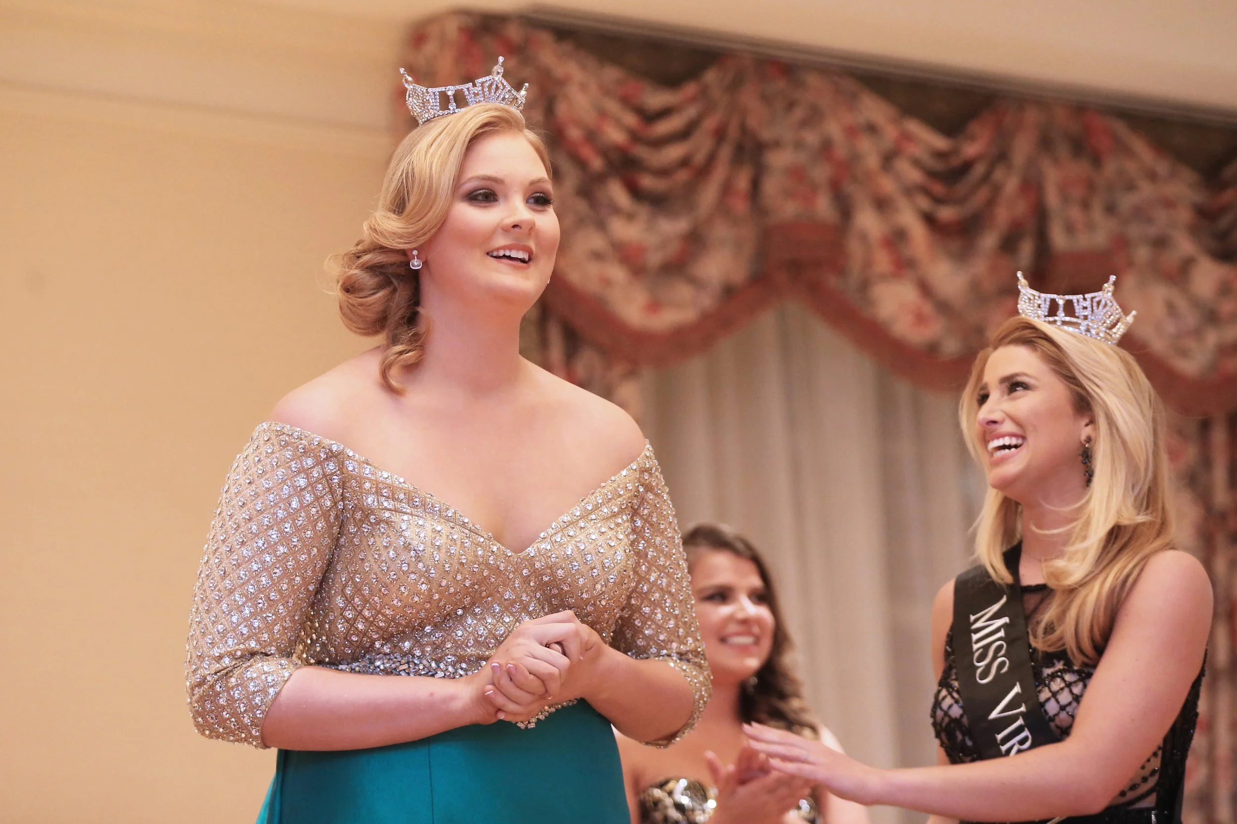 Miss Virginia wearing a crown and sparkling dress during a pageant, smiling with hands clasped, while being crowned by another woman with a sash that says Miss Virginia, in an elegant room with floral curtains.