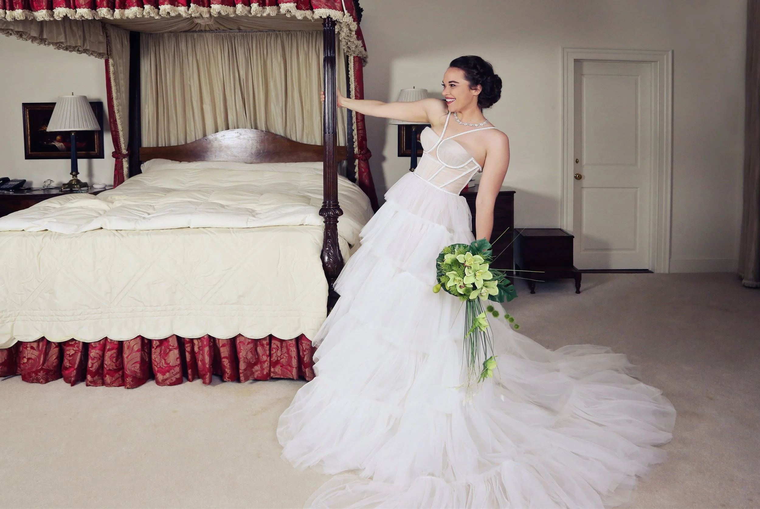 A woman in a wedding dress holding a bouquet, standing next to a four-poster bed in a hotel room.