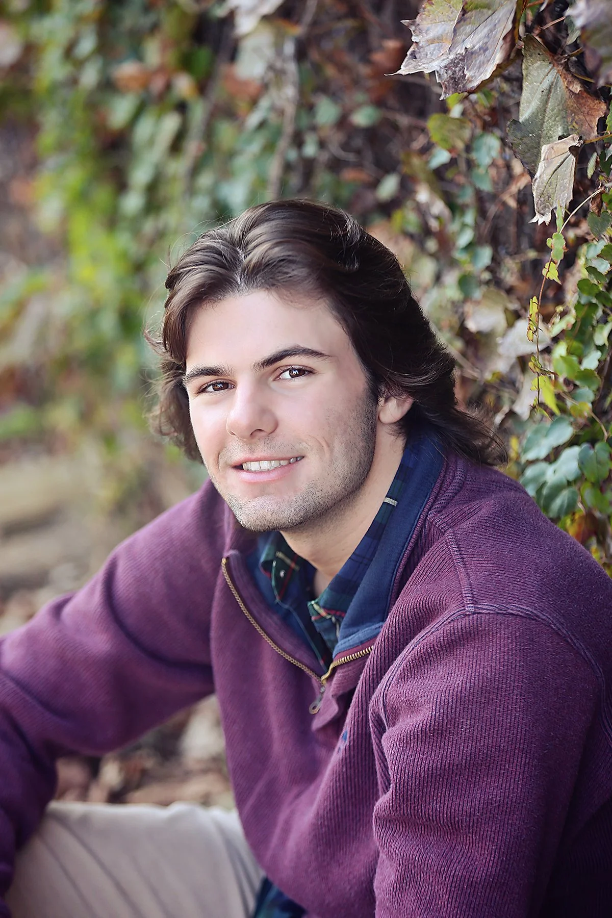 A young man with brown hair and a friendly smile, wearing a purple sweater over a plaid shirt, sitting outdoors near a leafy background.