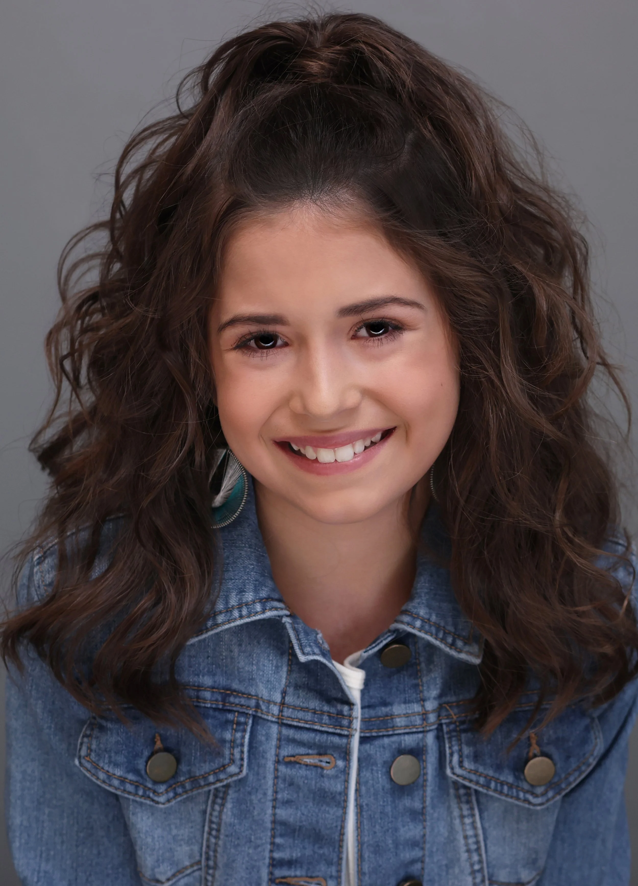 A young woman with wavy brown hair, smiling, wearing a denim jacket and silver earrings, against a neutral gray background.