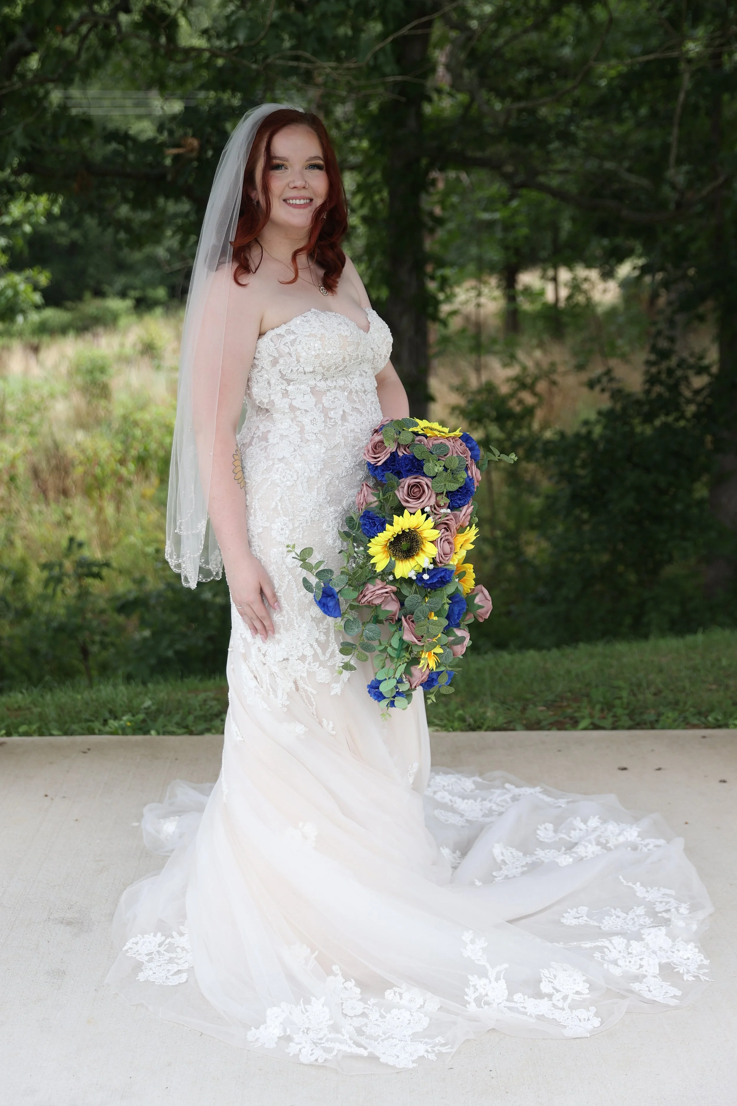 A bride in a strapless white wedding gown with lace details, holding a cascading bouquet of sunflowers, roses, and greenery, outdoors in a green, wooded area.