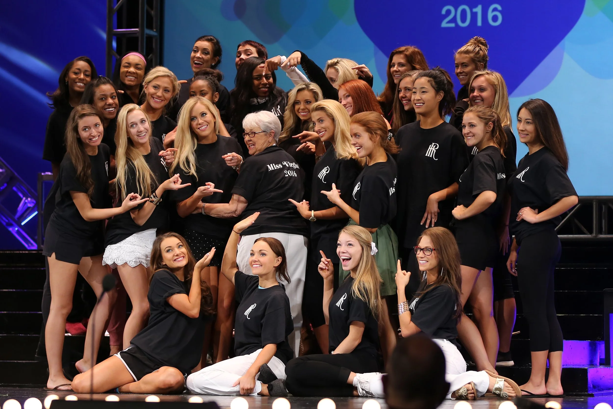 Group of women and young girls on stage at a beauty pageant event wearing black t-shirts with white text. An elderly woman with white hair and glasses is surrounded by the group. The background shows a large screen with the year "2016".