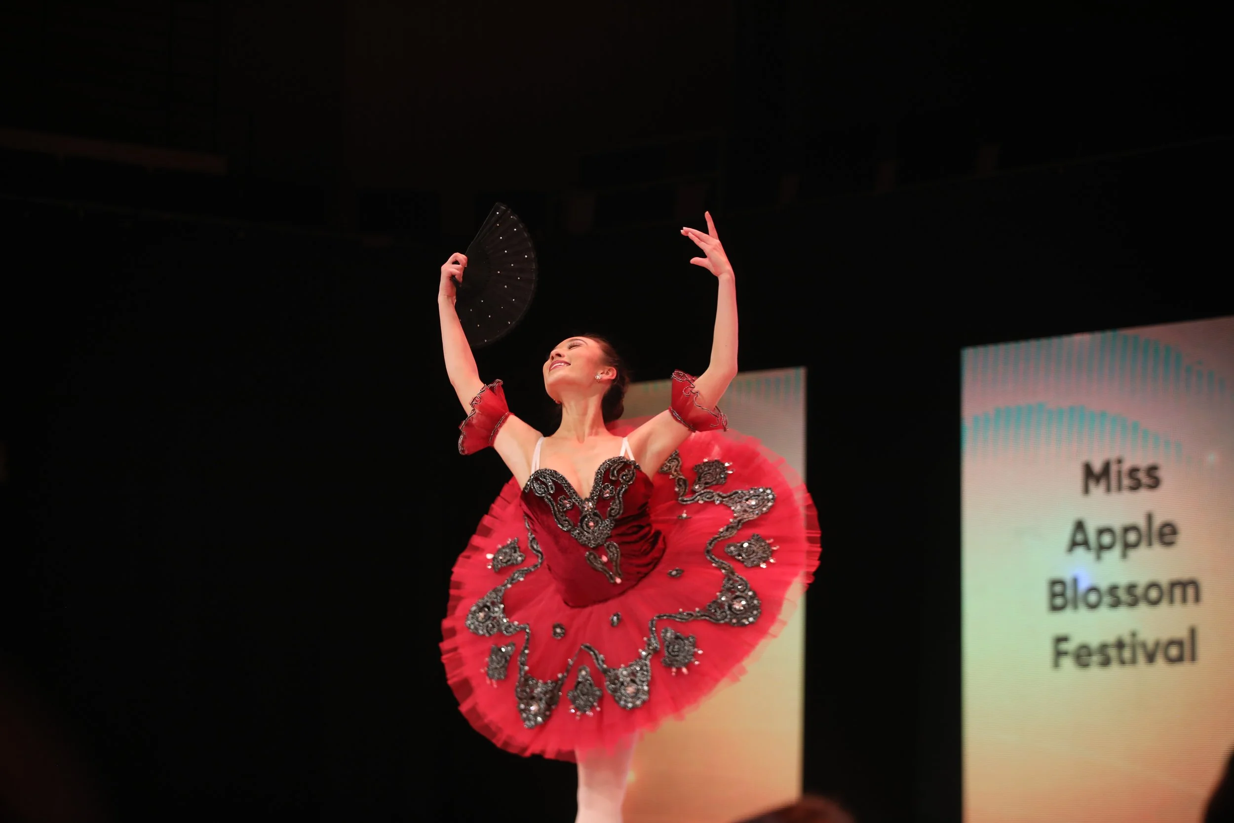 A woman in a red ballet costume with black and silver embellishments performing a dance on stage, holding a black fan, with a screen behind her that reads ‘Miss Apple Blossom Festival’.