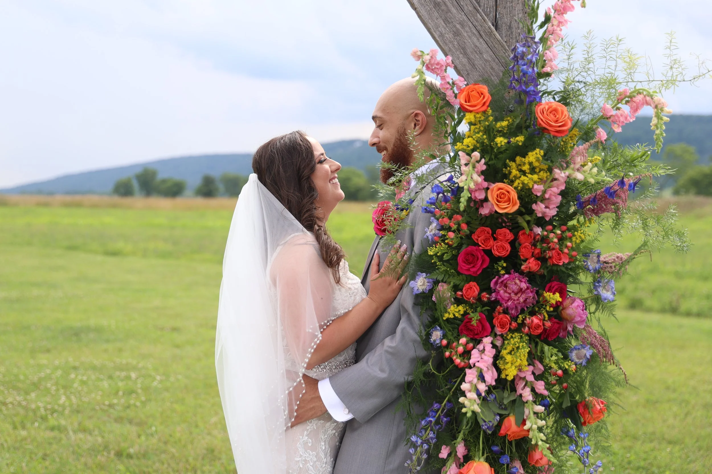A bride and groom holding each other and smiling at each other outdoors during their wedding in front of a large, colorful floral arrangement on a wooden post, with a grassy field and hills in the background.