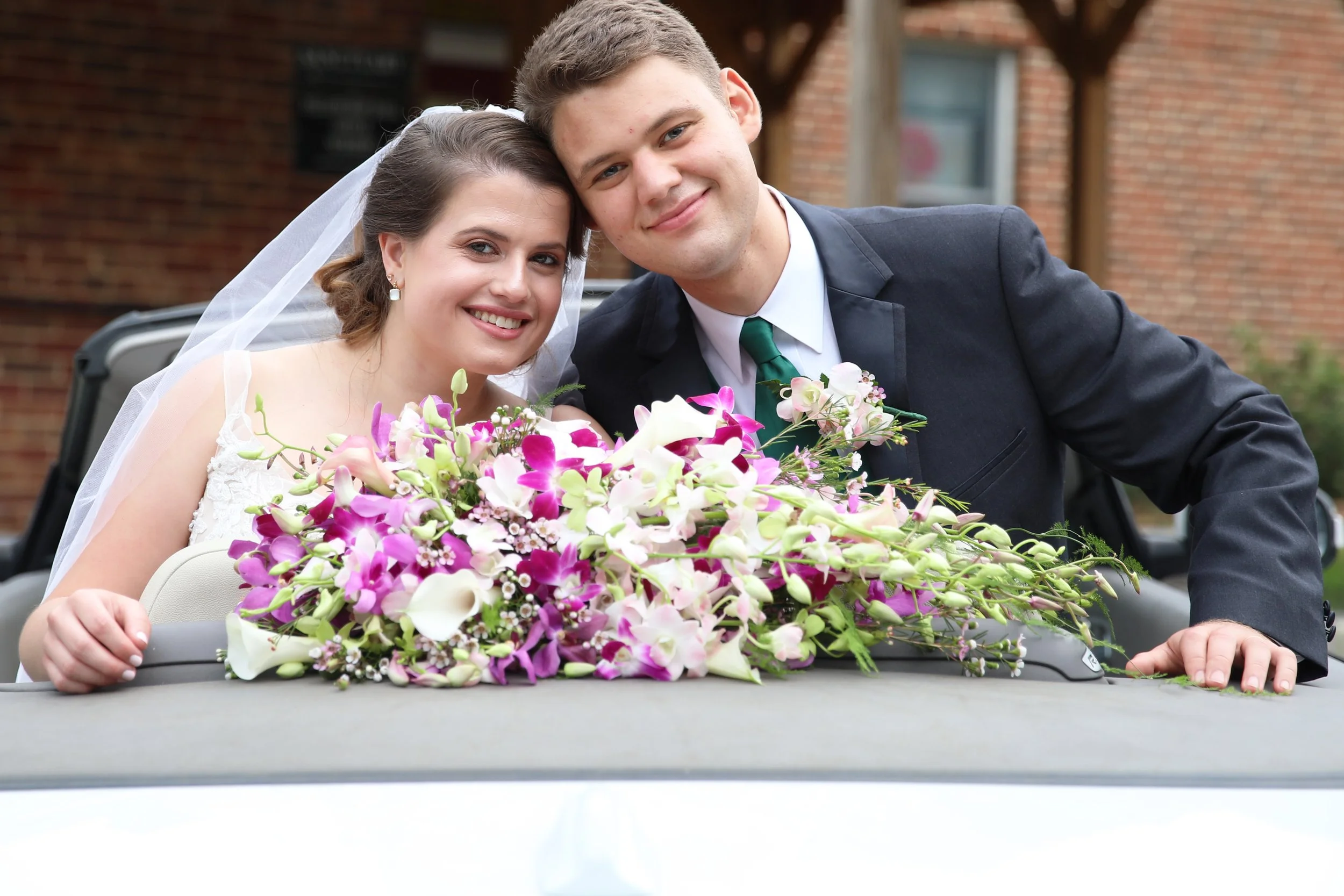 Smiling bride and groom leaning on a car decorated with a large bouquet of pink, white, and purple flowers during a wedding celebration.