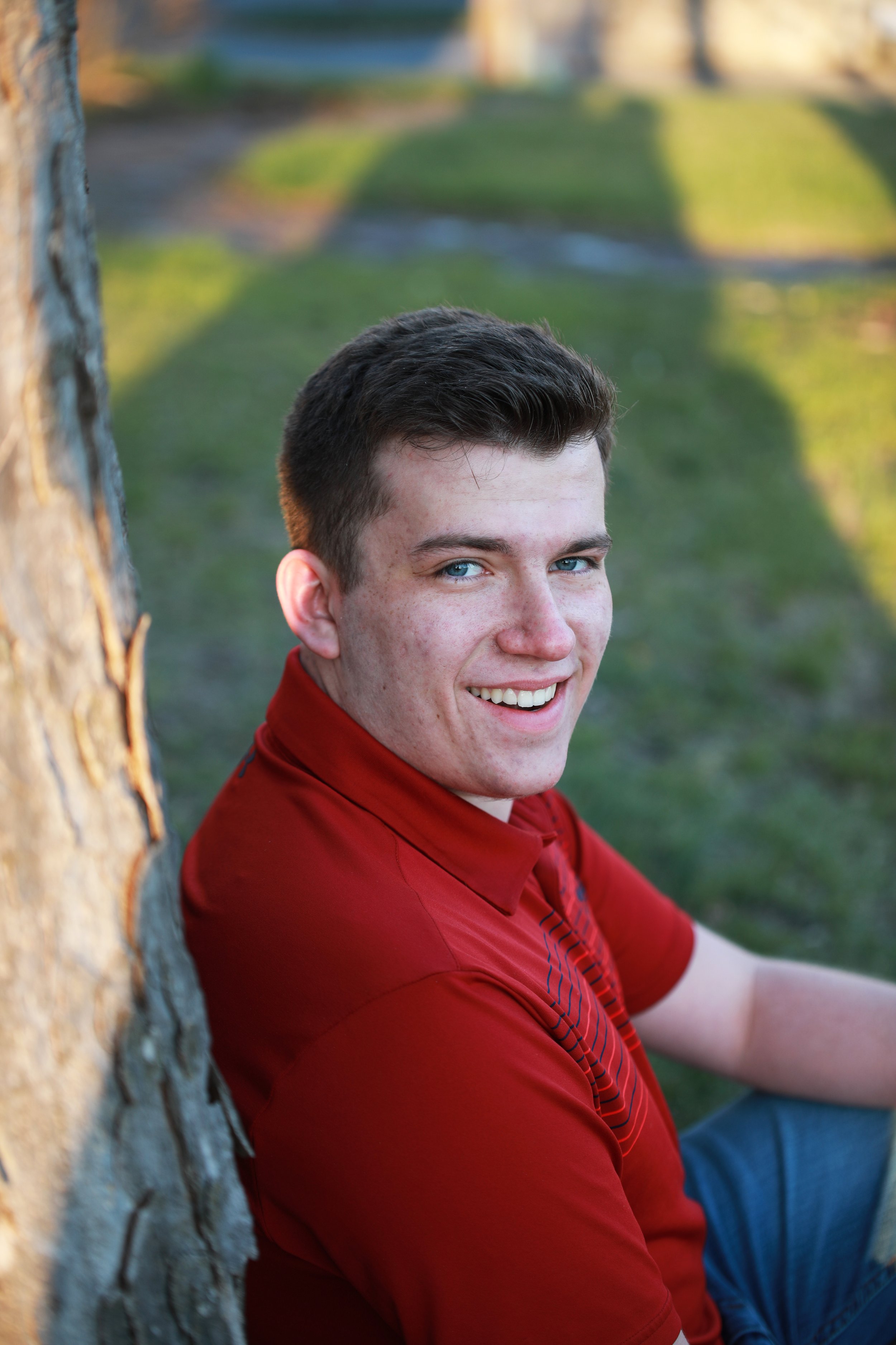 A young man with short brown hair and blue eyes sitting outdoors on grass, leaning against a tree, smiling at the camera in late afternoon sunlight, wearing a red shirt.