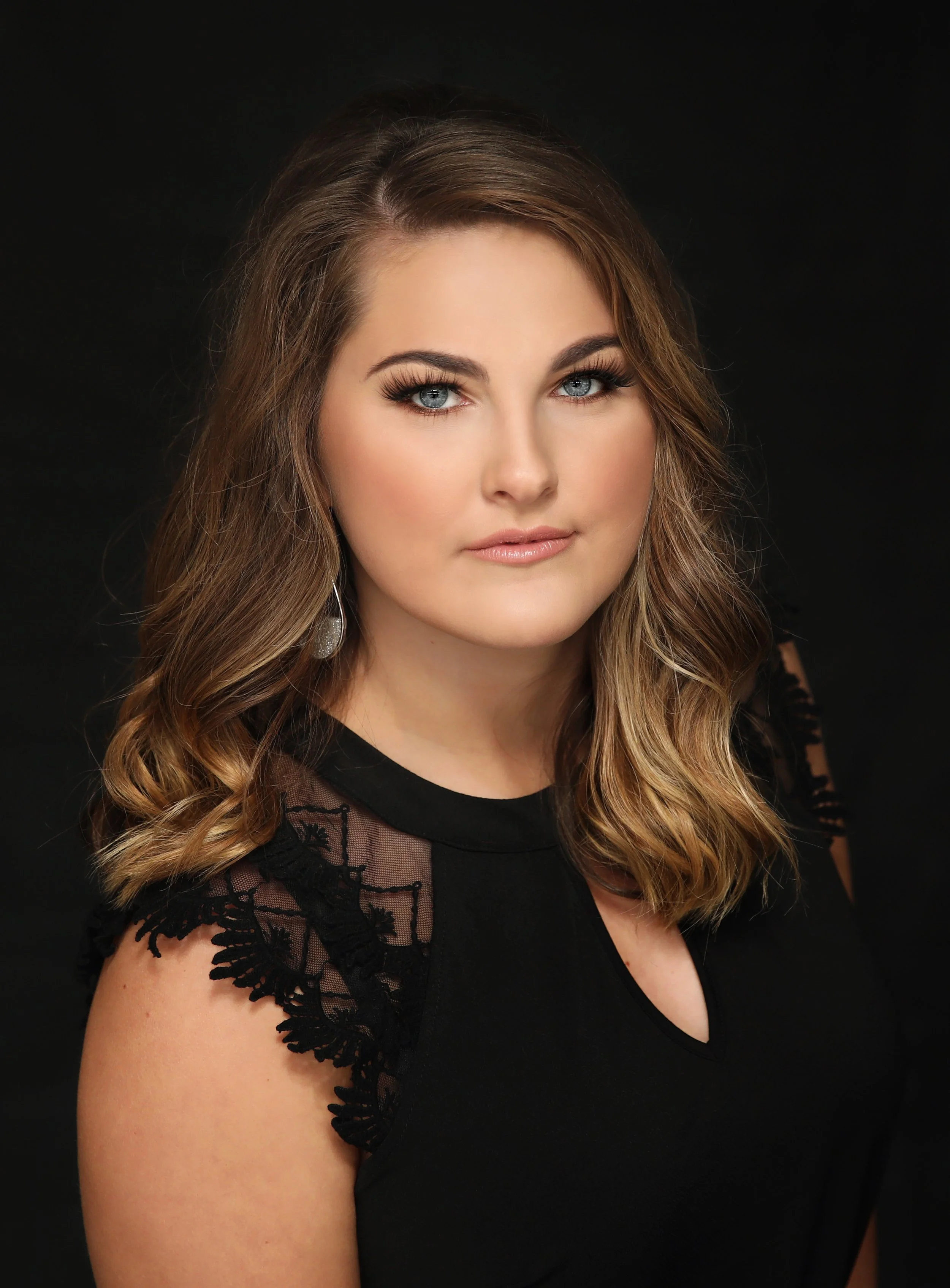 Portrait of a woman with wavy brown hair, blue eyes, and makeup, wearing a black top with lace details, against a dark background.