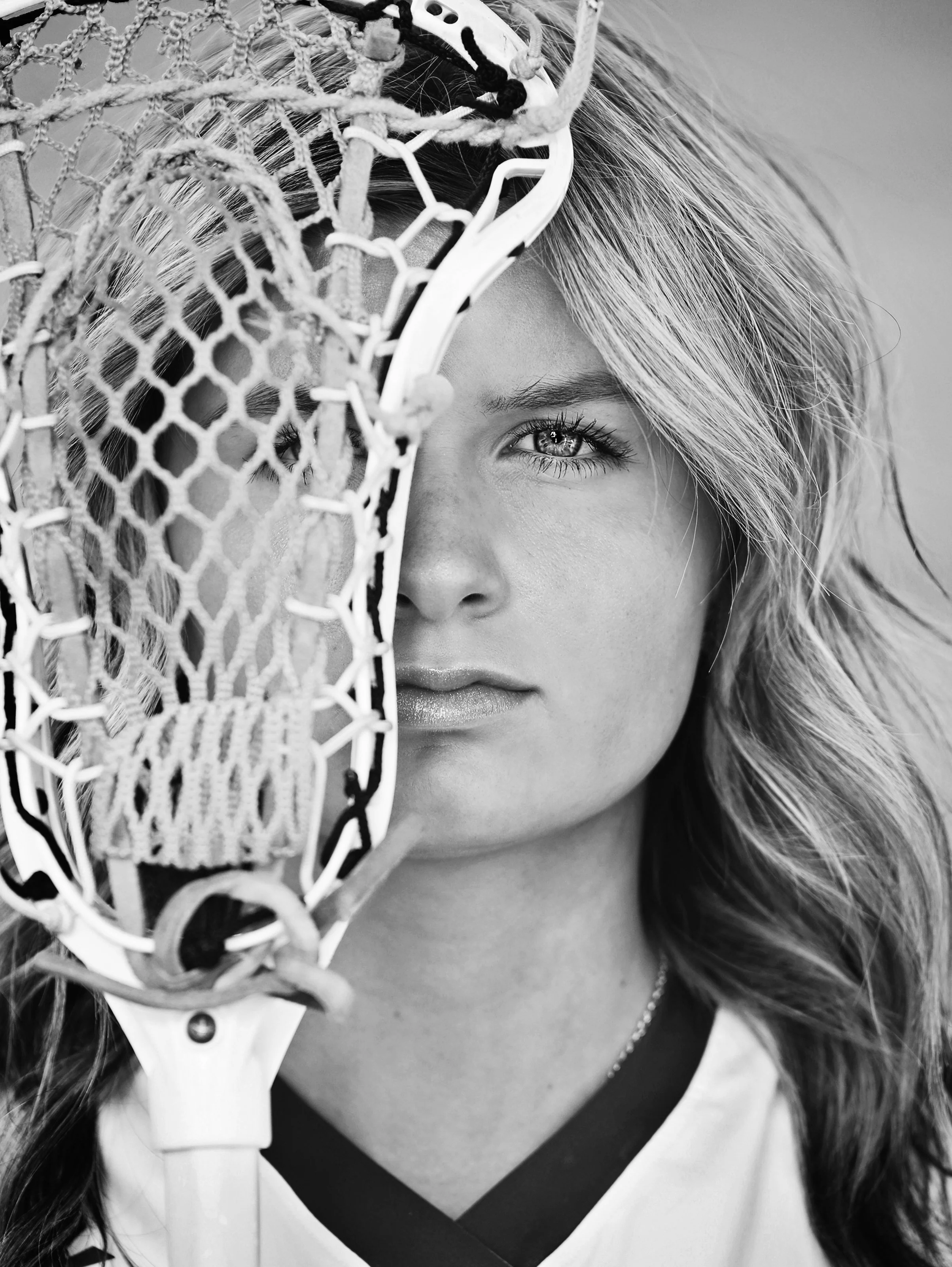 Close-up of a woman's face partially obscured by a lacrosse stick with a netted scoop, focusing on her eye and facial features, black and white photograph.
