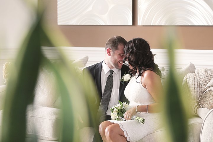A couple, dressed in wedding attire, sitting close together on a beige couch, smiling and leaning in for a kiss, with greenery partially obscuring the view.