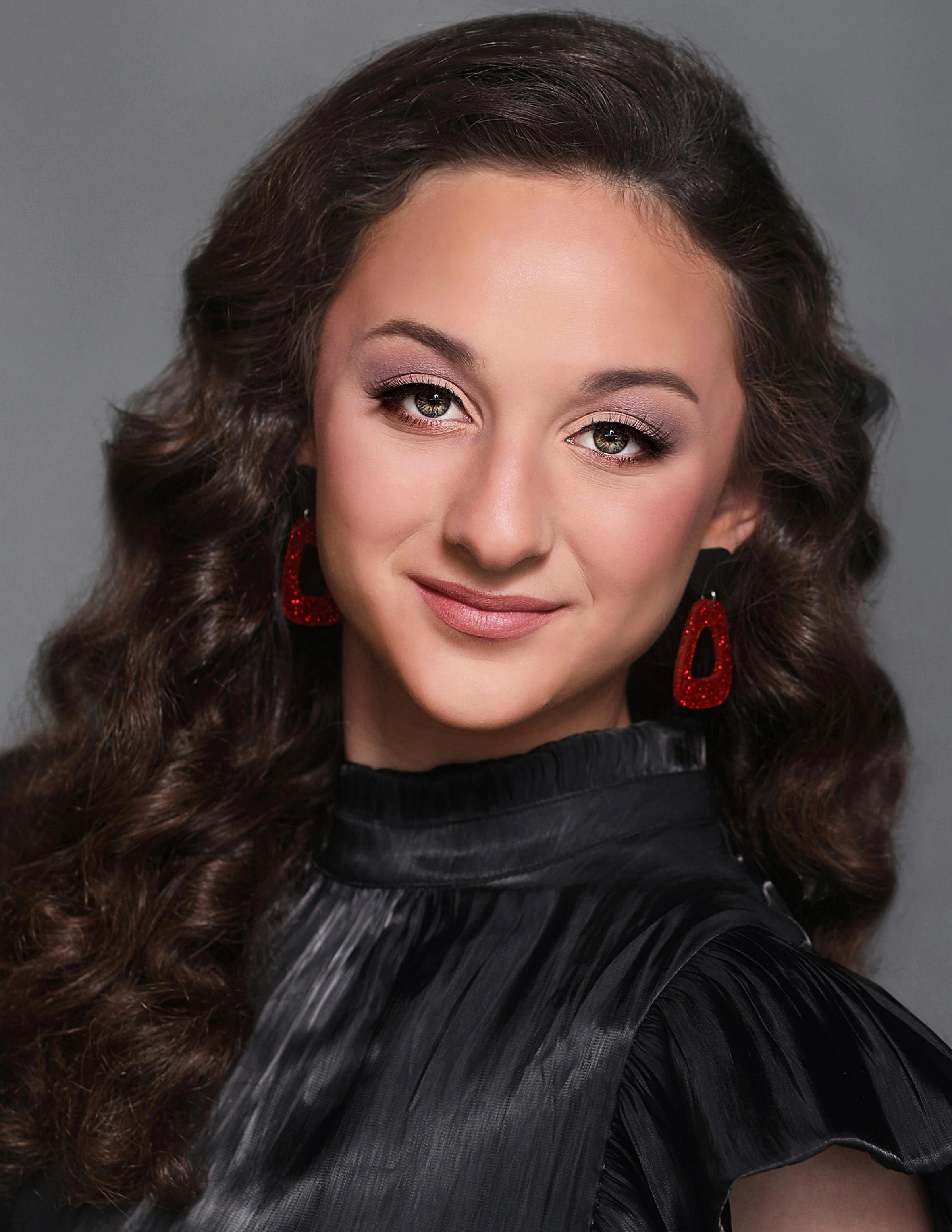 Close-up portrait of a woman with long, curly brown hair wearing red earrings and a black shiny top, smiling softly against a gray background.