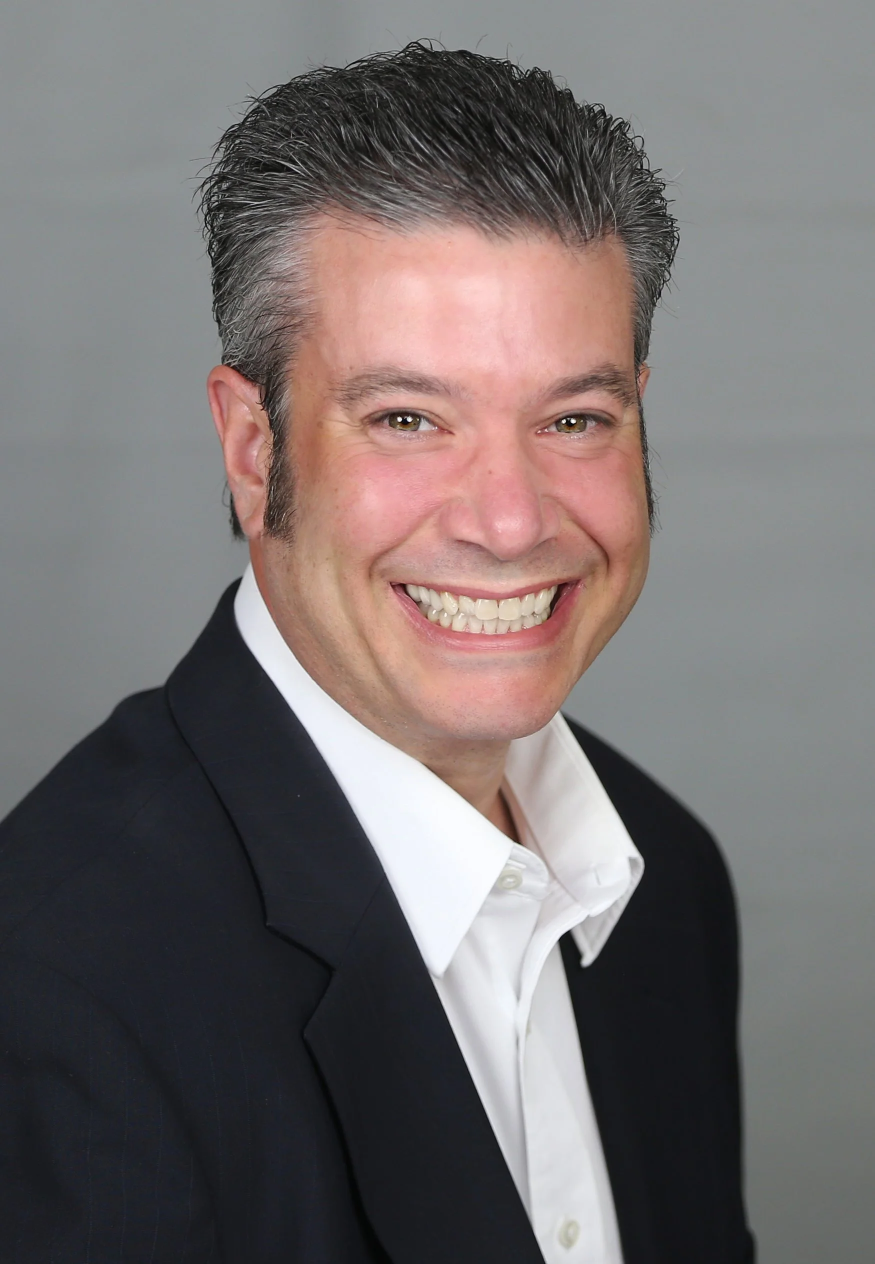 Headshot of a smiling man with dark, slicked-back hair, wearing a black suit and white shirt, against a gray background.