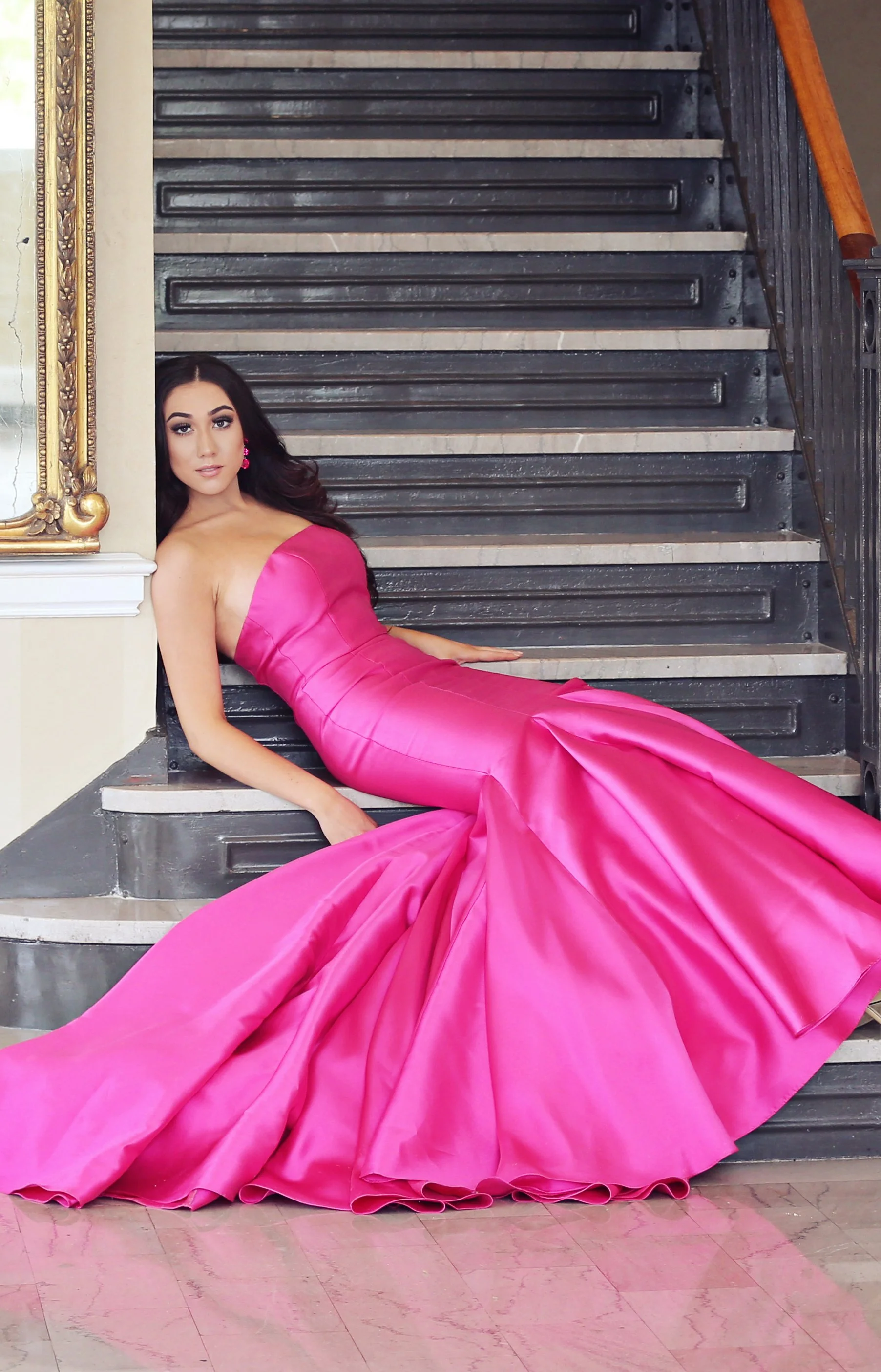 A woman in a strapless bright pink satin gown sitting on a staircase with a black and white tiled step, posing for the camera inside a building with a gold-framed mirror, a white wall, and wooden railings.