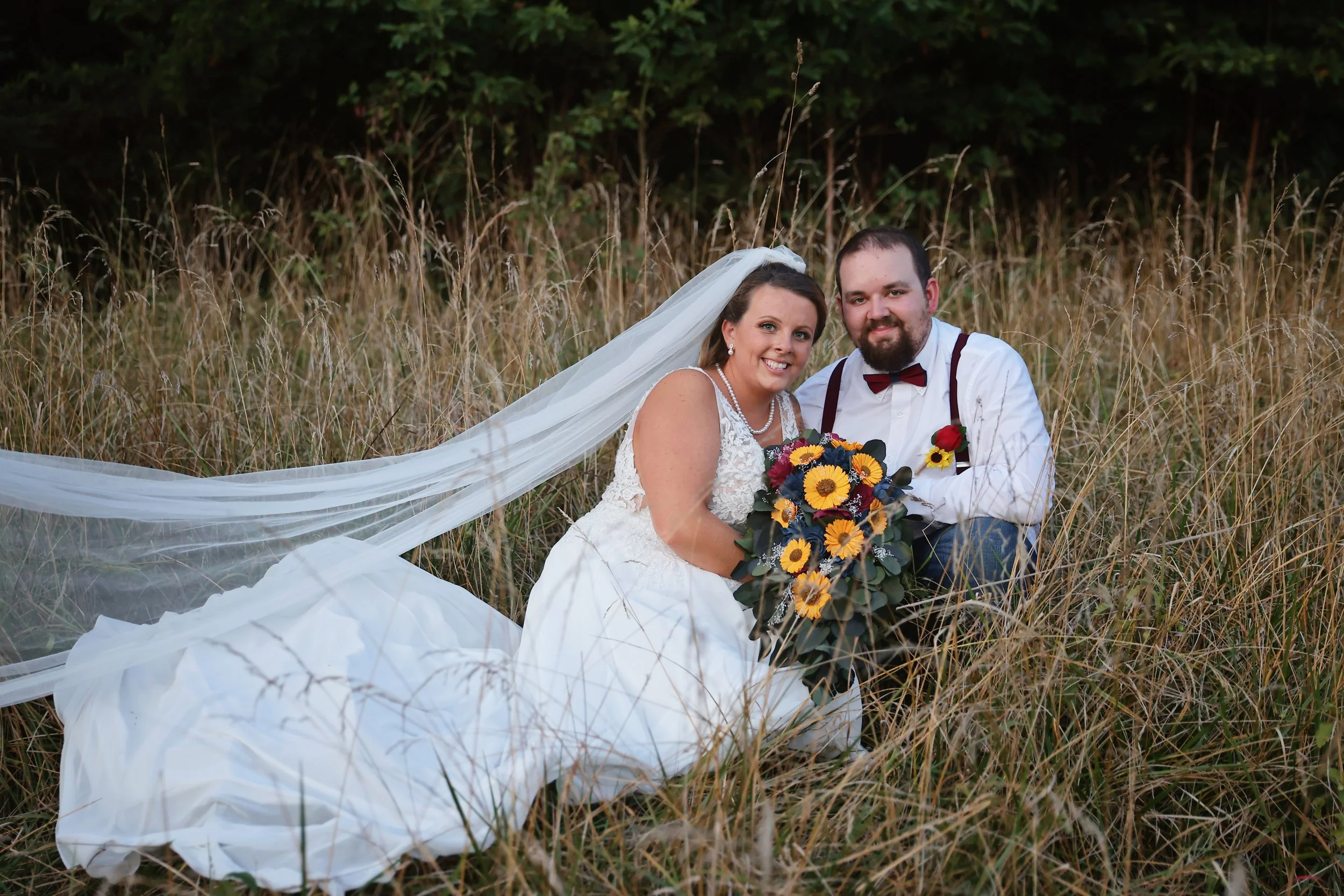 A newlywed couple sitting in a field of tall grass, smiling at the camera. The bride wears a white wedding dress with a long veil and holds a bouquet of sunflowers and other flowers. The groom wears a white shirt with suspenders, a burgundy bow tie, and has a boutonniere on his shirt.