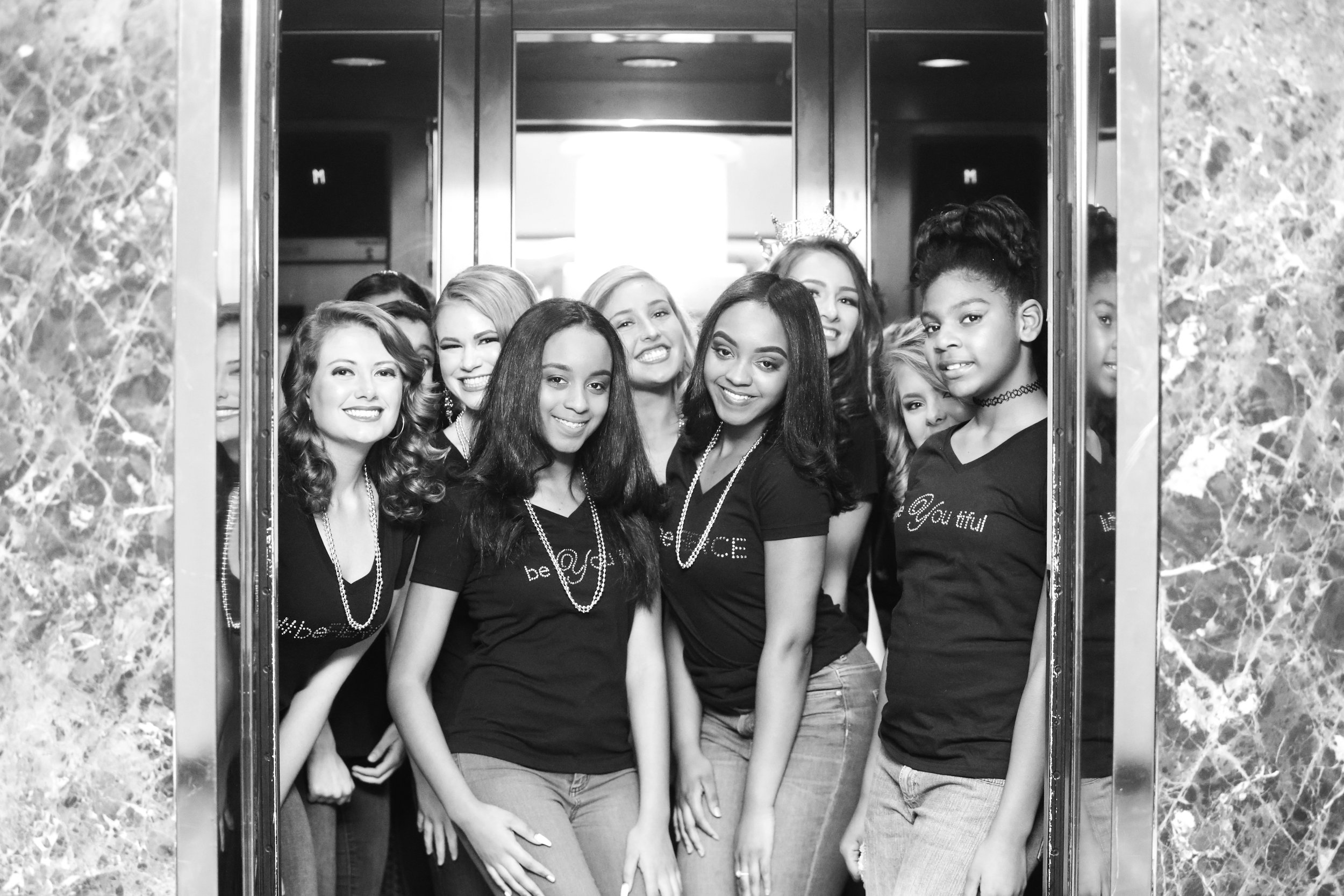 Group of women and girls inside an elevator, smiling for a photo. They are wearing matching T-shirts that say 'be YOUtiful' and accessories like necklaces and a crown. The setting appears to be a celebration or event.