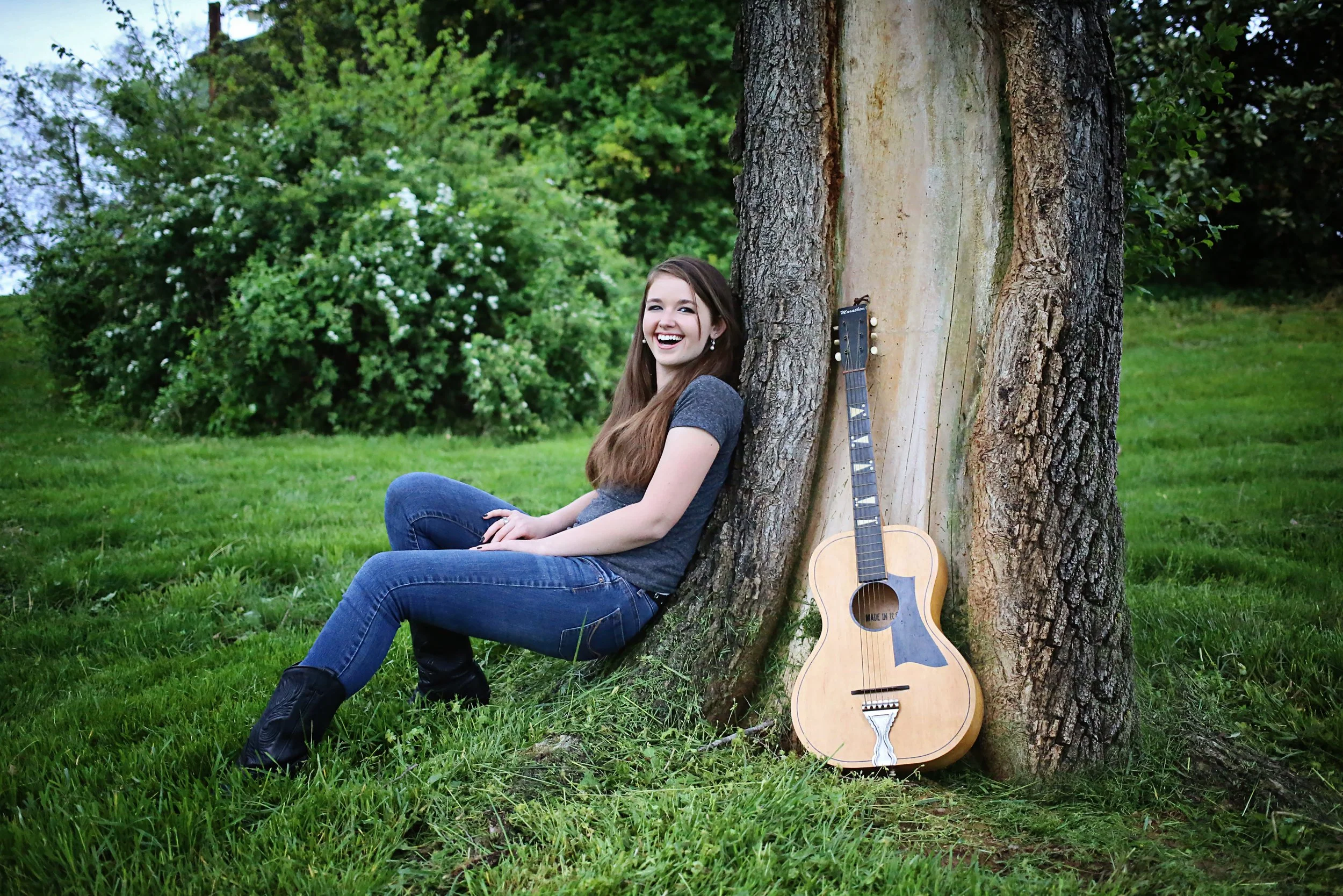A young woman with long brown hair, wearing a dark gray t-shirt and jeans, sitting on grass and leaning against a large tree, smiling at the camera. An acoustic guitar rests against the tree next to her in a park-like setting with green grass, trees, and bushes in the background.