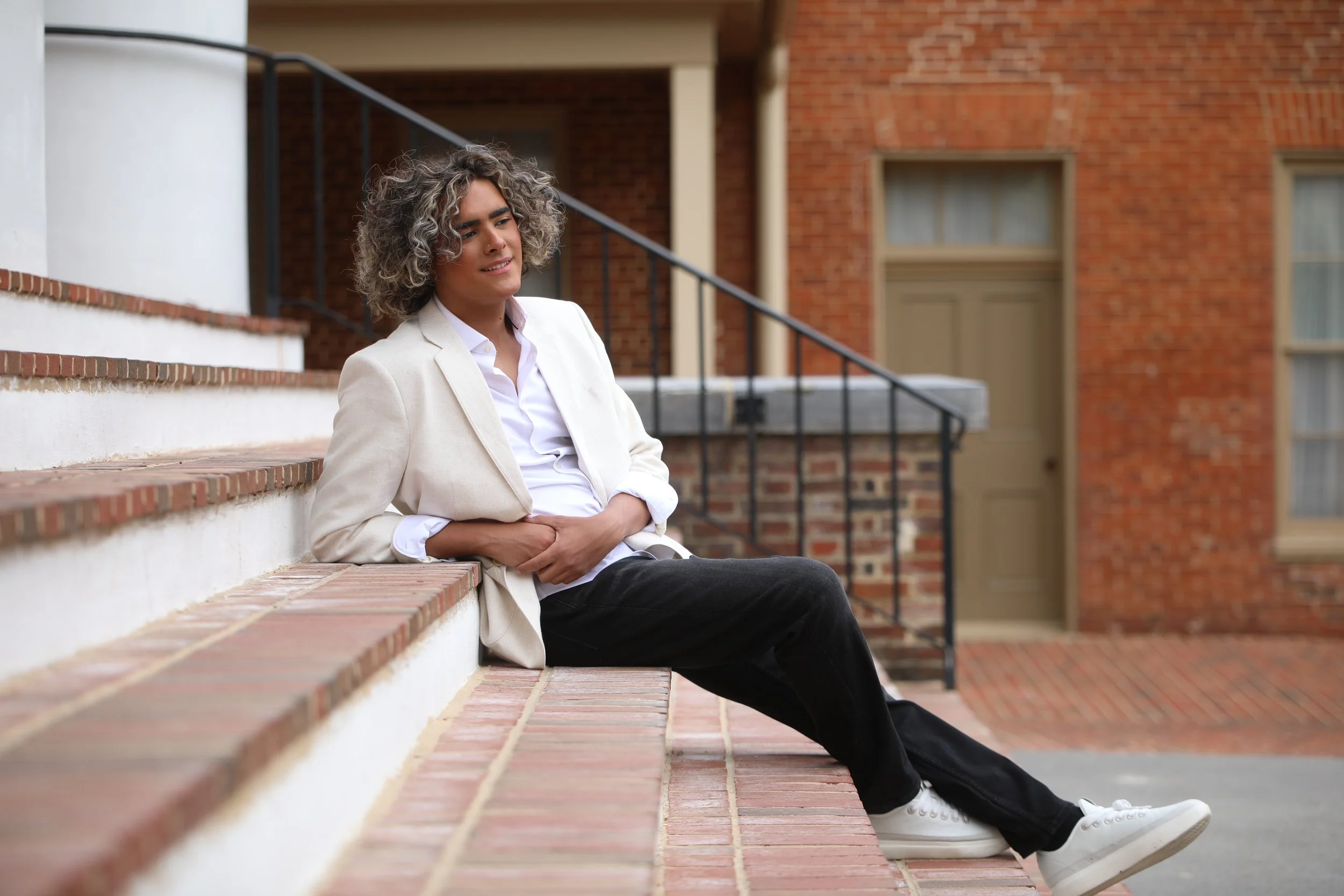 A young person with curly hair sitting on brick steps outside a brick building, wearing a white shirt, light-colored blazer, black pants, and white sneakers.