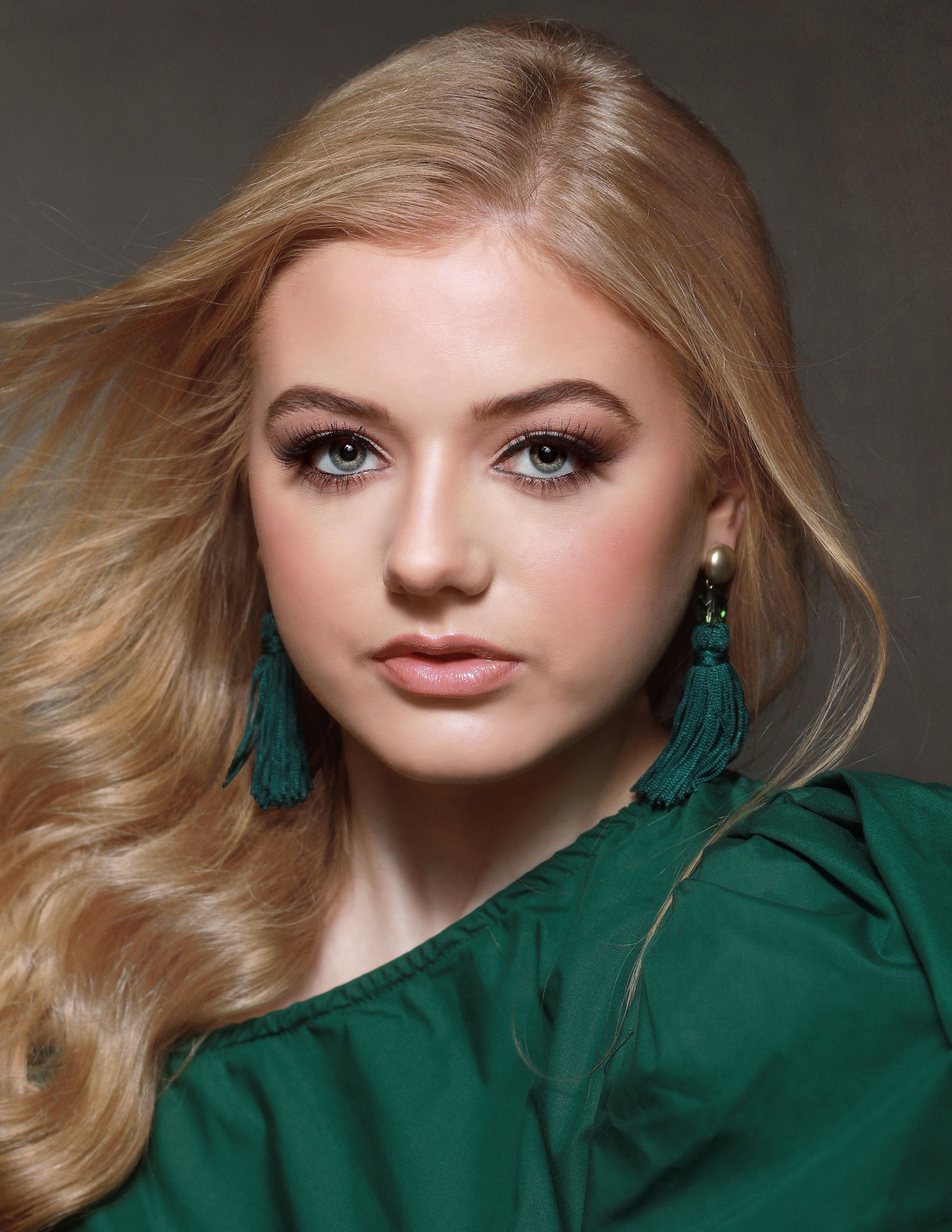 Close-up portrait of a young woman with long blonde hair, wearing dark eye makeup, peach lipstick, green earrings with tassels, and a green blouse against a gray background.