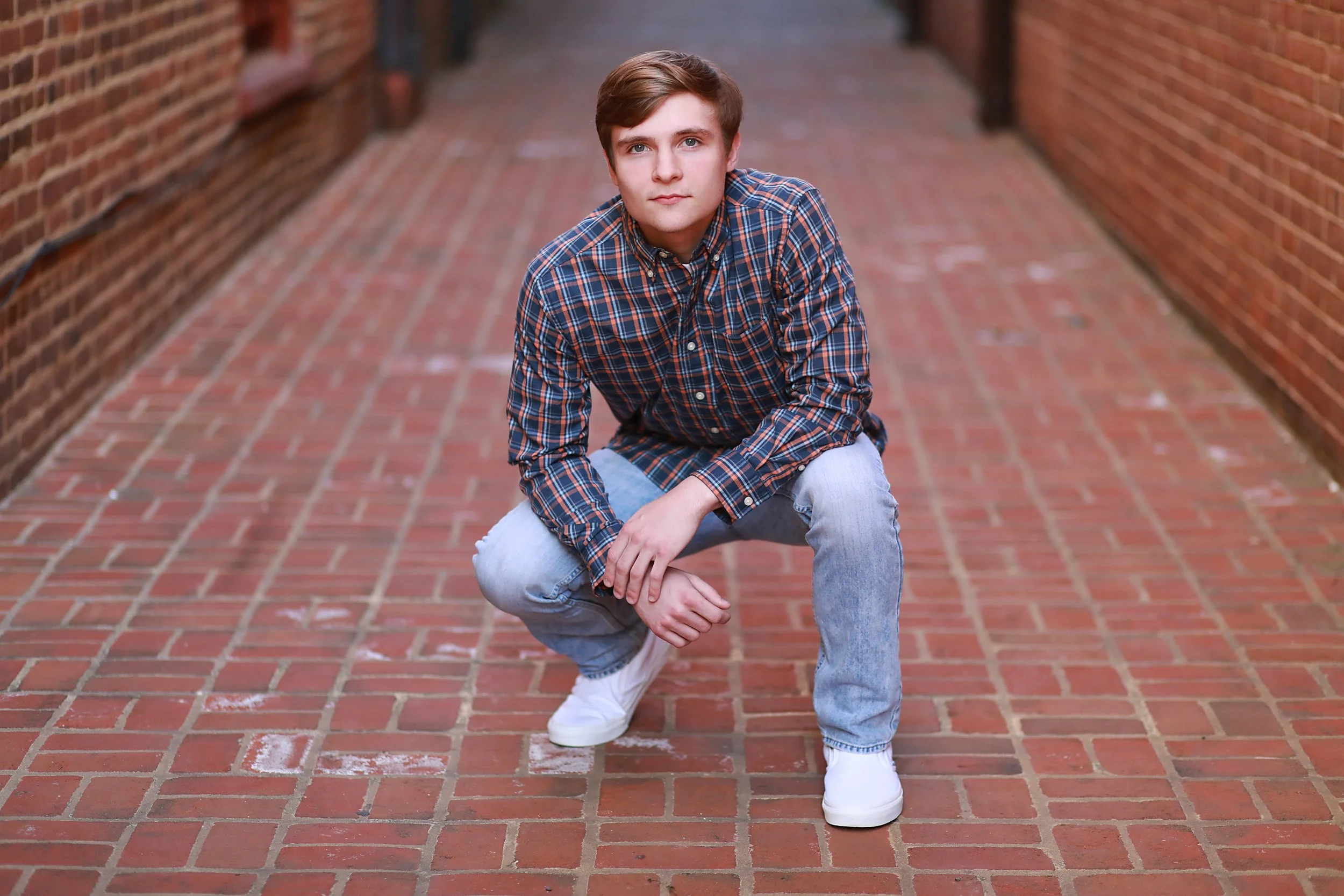 Young man in plaid shirt and jeans crouching on a brick alleyway, looking at the camera.