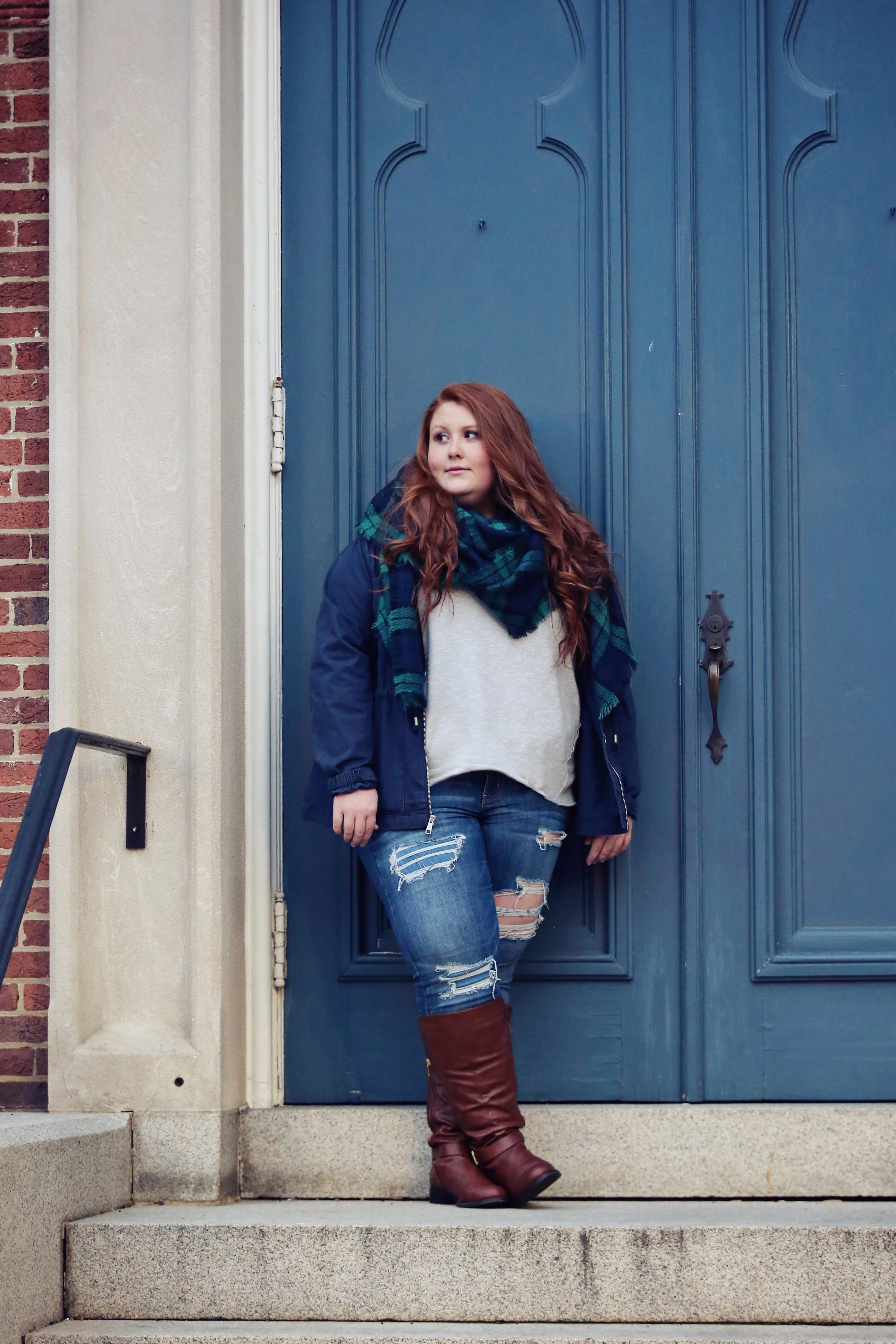 A young woman with long red hair leans against a blue door, wearing a navy jacket, plaid scarf, gray sweater, ripped jeans, and brown boots, standing on concrete steps outside a building.