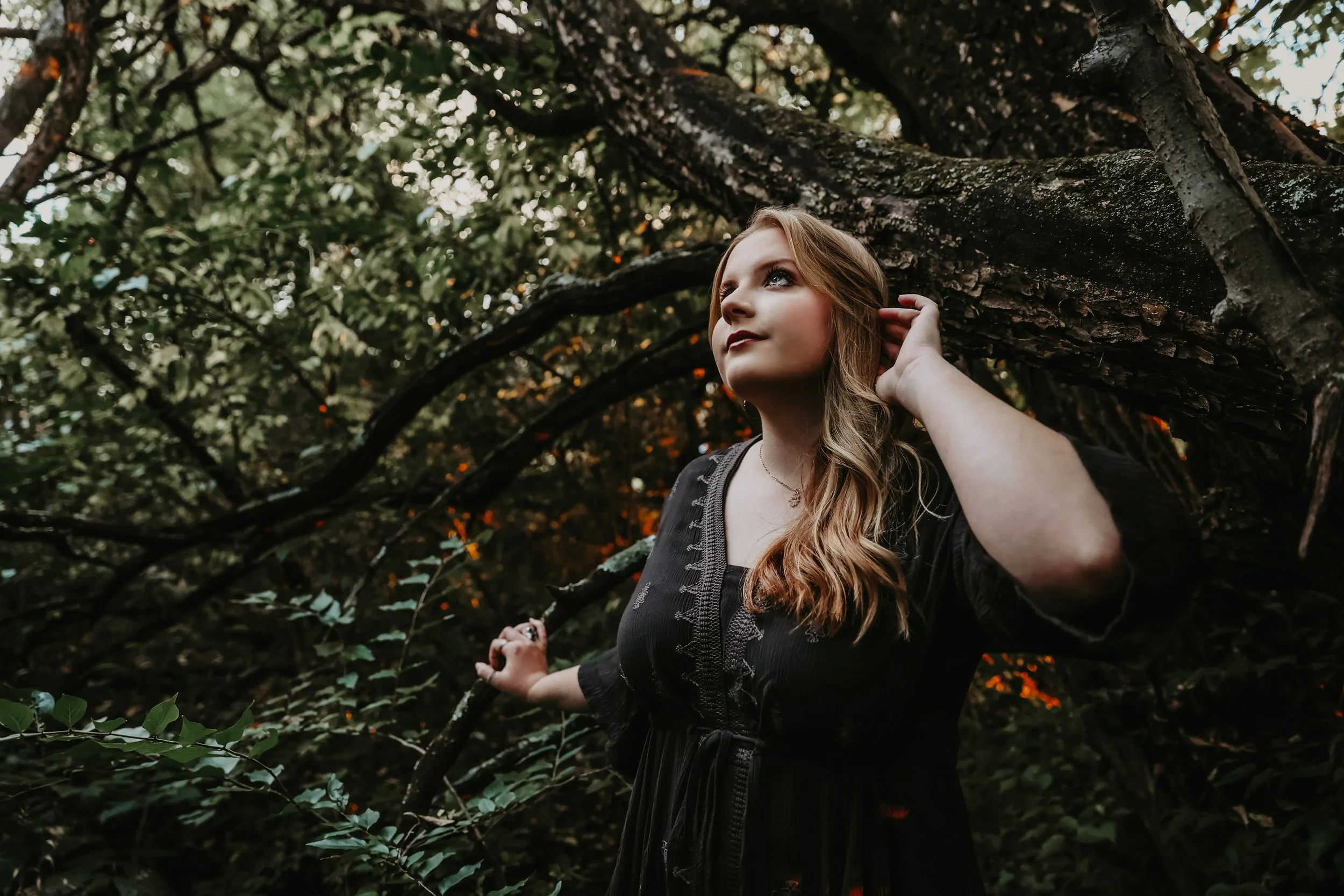 Young woman with long wavy hair standing outdoors in a wooded area, touching a tree branch behind her head and looking upward.