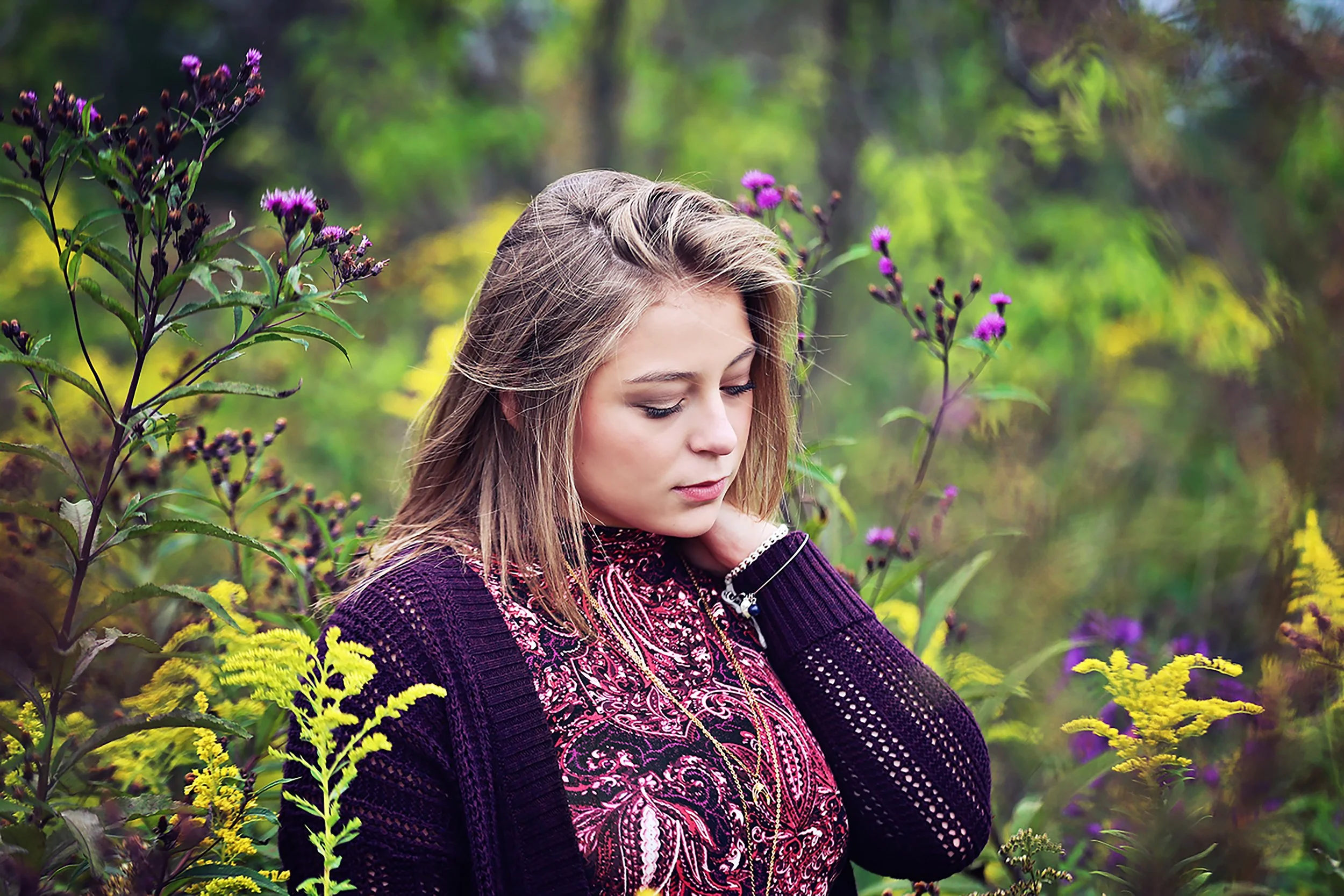 A young woman with blonde hair, wearing a purple and red patterned top and a black open-knit sweater, standing among purple and yellow flowers in a green outdoor setting, looking down with her hand touching her neck.