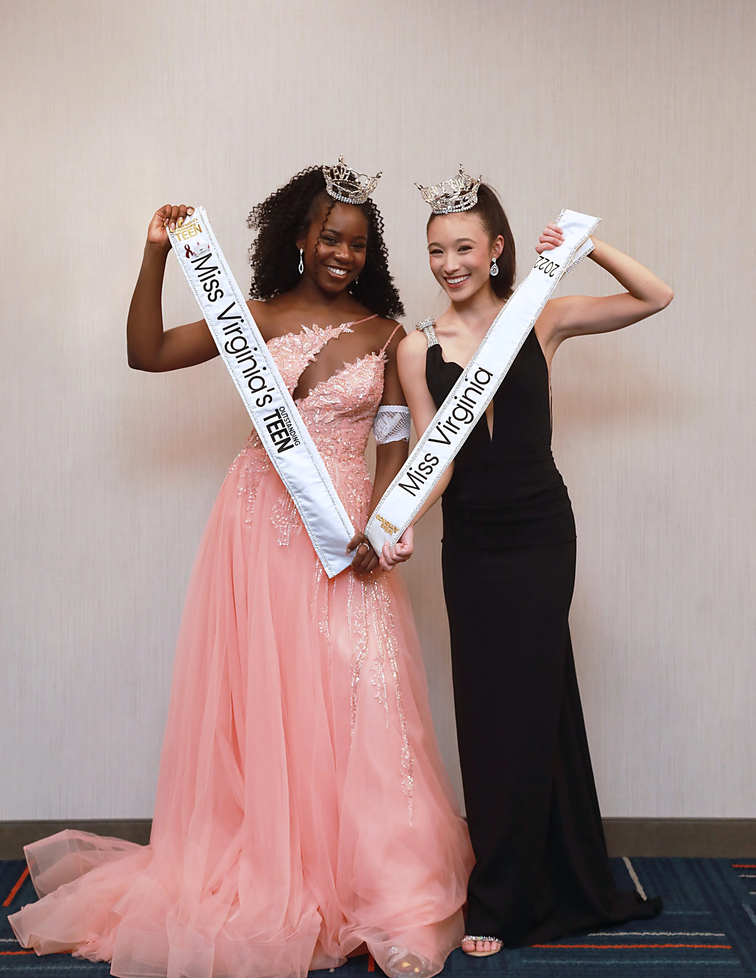 Two women wearing crowns and sashes that read "Miss Virginia" and "Miss Virginia's Teen" stand together, celebrating their pageant wins. One is in a pink gown, and the other in a black gown.