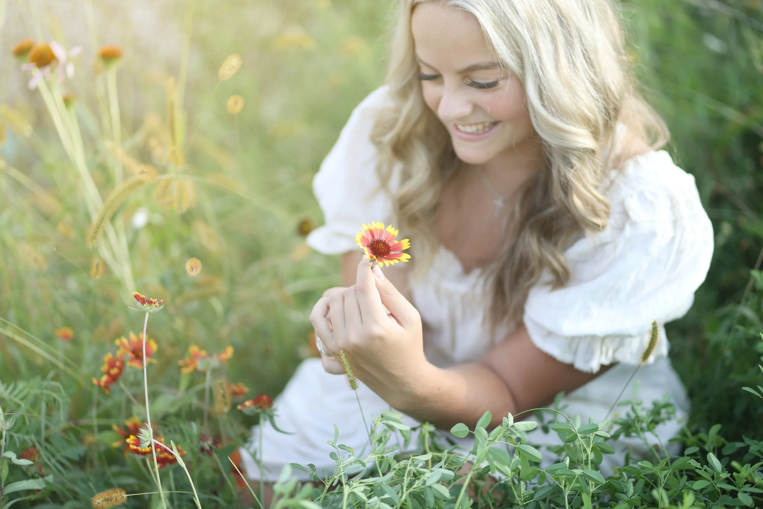 A woman with long blonde hair in a white dress smiling while holding a yellow and red flower in a grassy field.