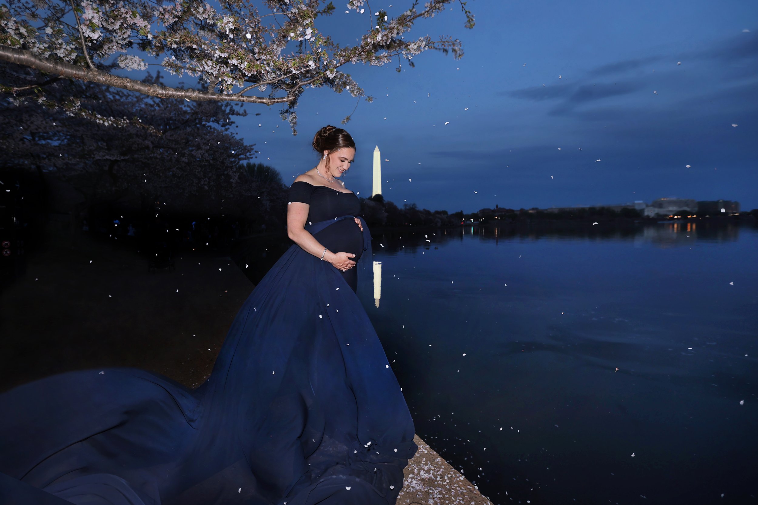 A pregnant woman wearing a navy blue off-shoulder dress standing by the water's edge during the evening, with cherry blossom trees overhead and the Washington Monument in the background, while petals or snowflakes fall around her.
