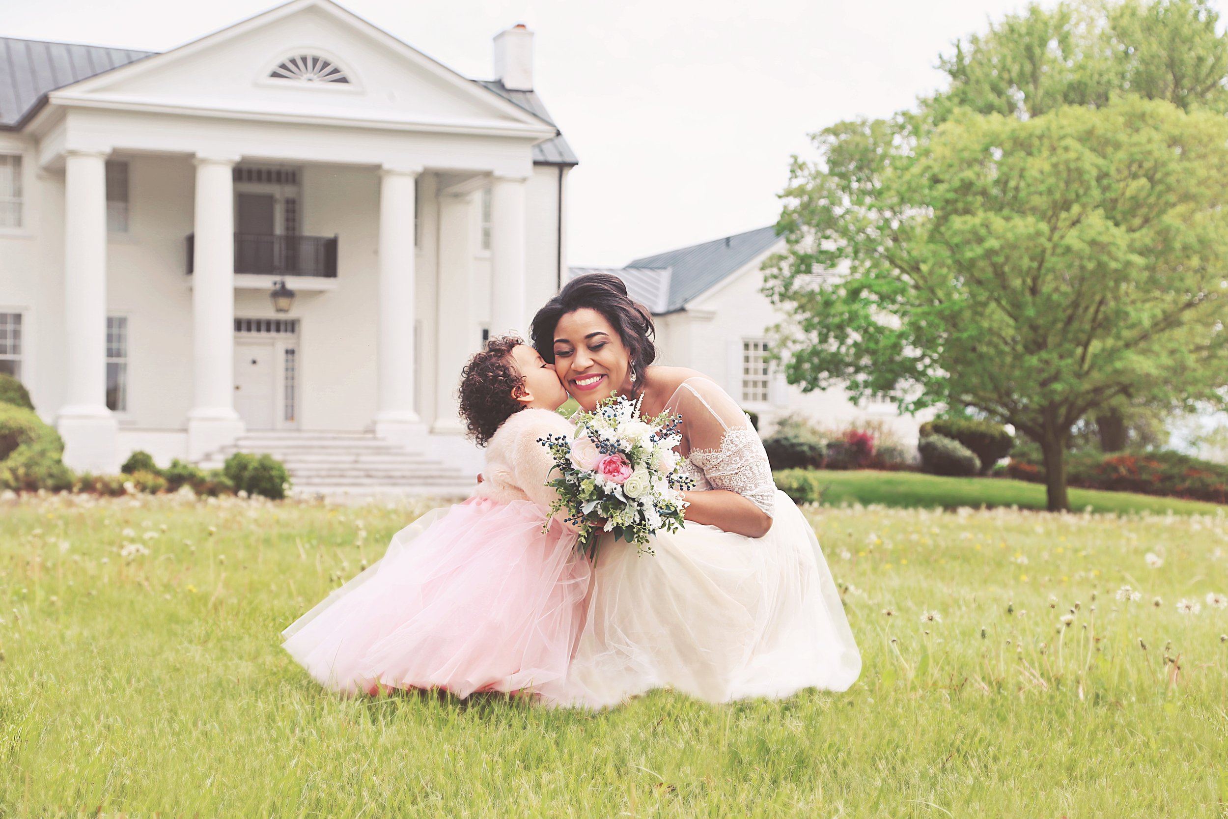 A woman in a white dress holding a bouquet of flowers receives a kiss from a young girl in a pink dress on a grassy lawn in front of a large white house with columns and a tree.