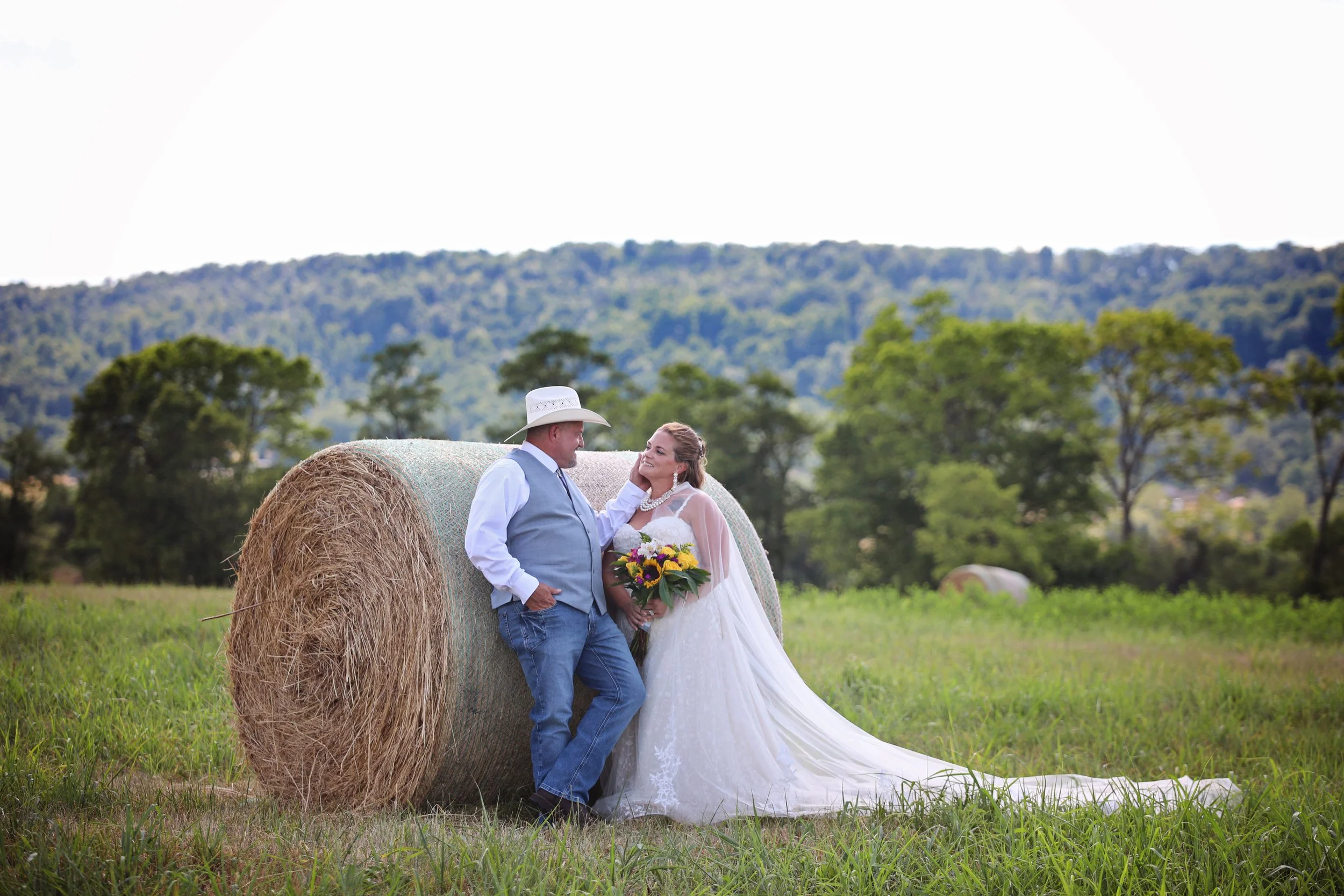 A wedding couple in a field with green grass and trees, standing next to a large hay bale, with mountain hills in the background. The bride is holding a bouquet, and the groom is wearing a cowboy hat.