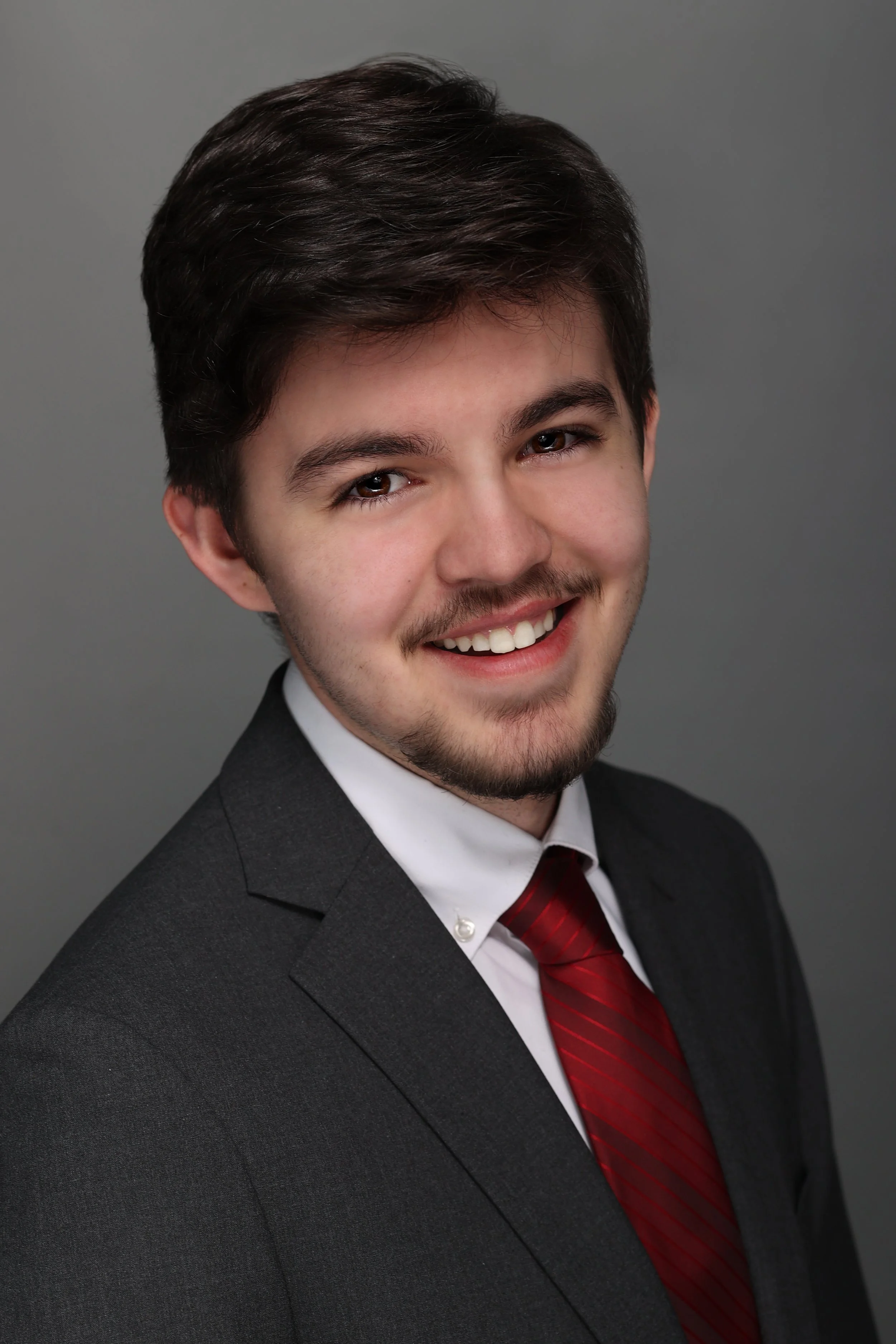 Young man with dark hair, beard, and mustache smiling, dressed in a suit and red tie against a gray background.