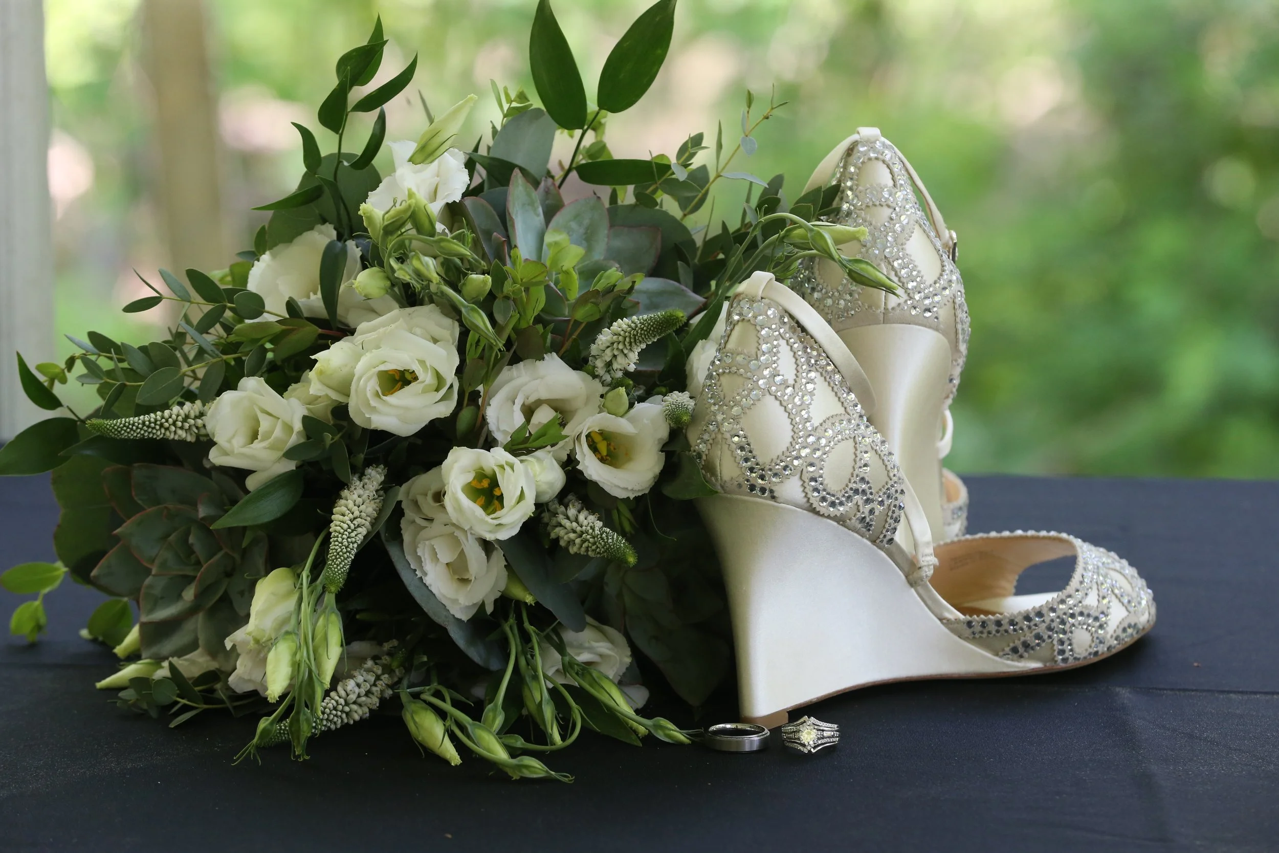 A bouquet of white flowers and greenery, a pair of white high-heeled shoes with intricate rhinestone detailing, a silver wedding band, and a star-shaped engagement ring on a black surface with a blurred green outdoor background.