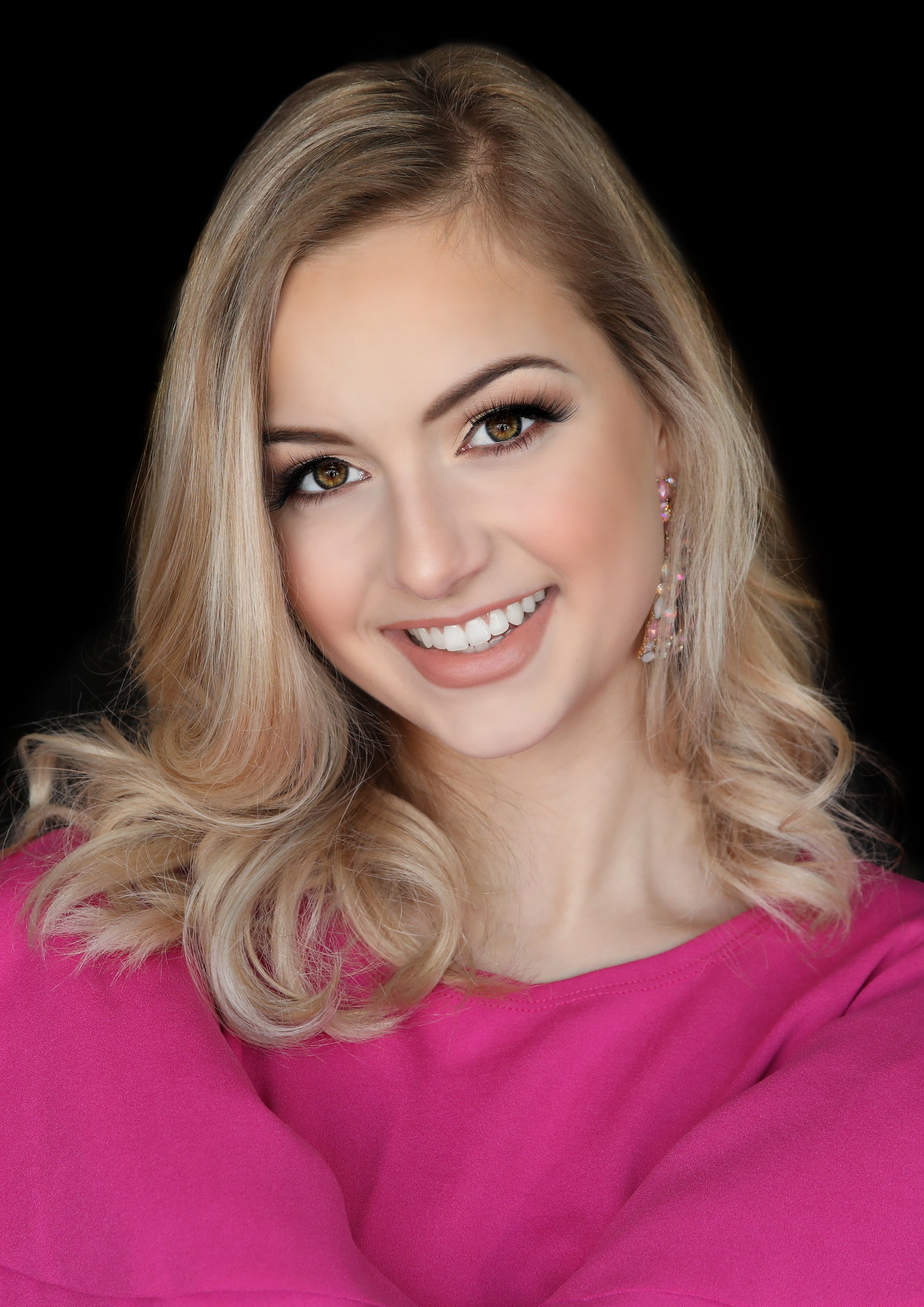 Close-up portrait of a smiling woman with blonde wavy hair, wearing pink earrings and a pink top, against a black background.