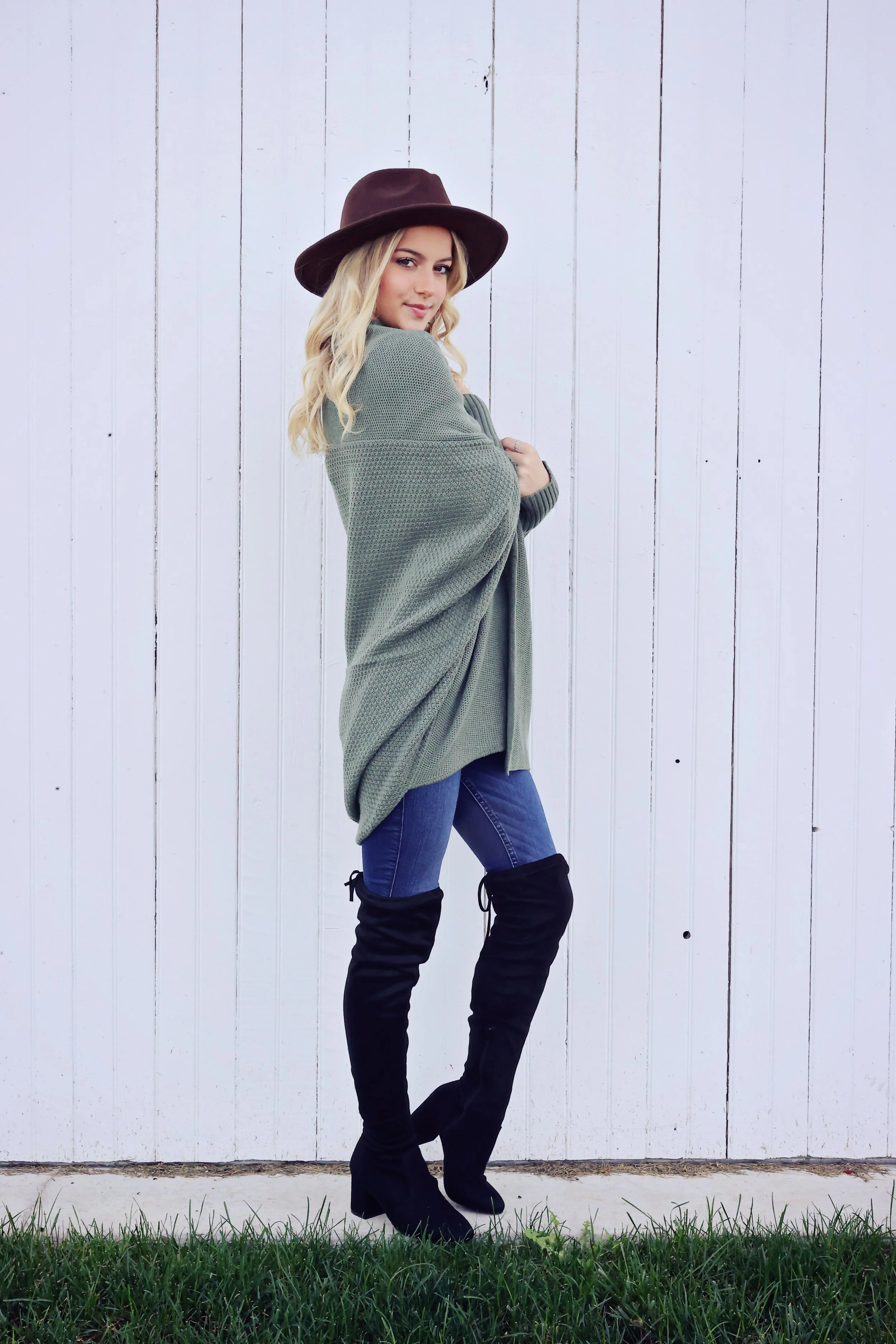 A young woman with blonde hair wearing a brown hat, an oversized green sweater, jeans, and black thigh-high boots standing against a white wooden wall.