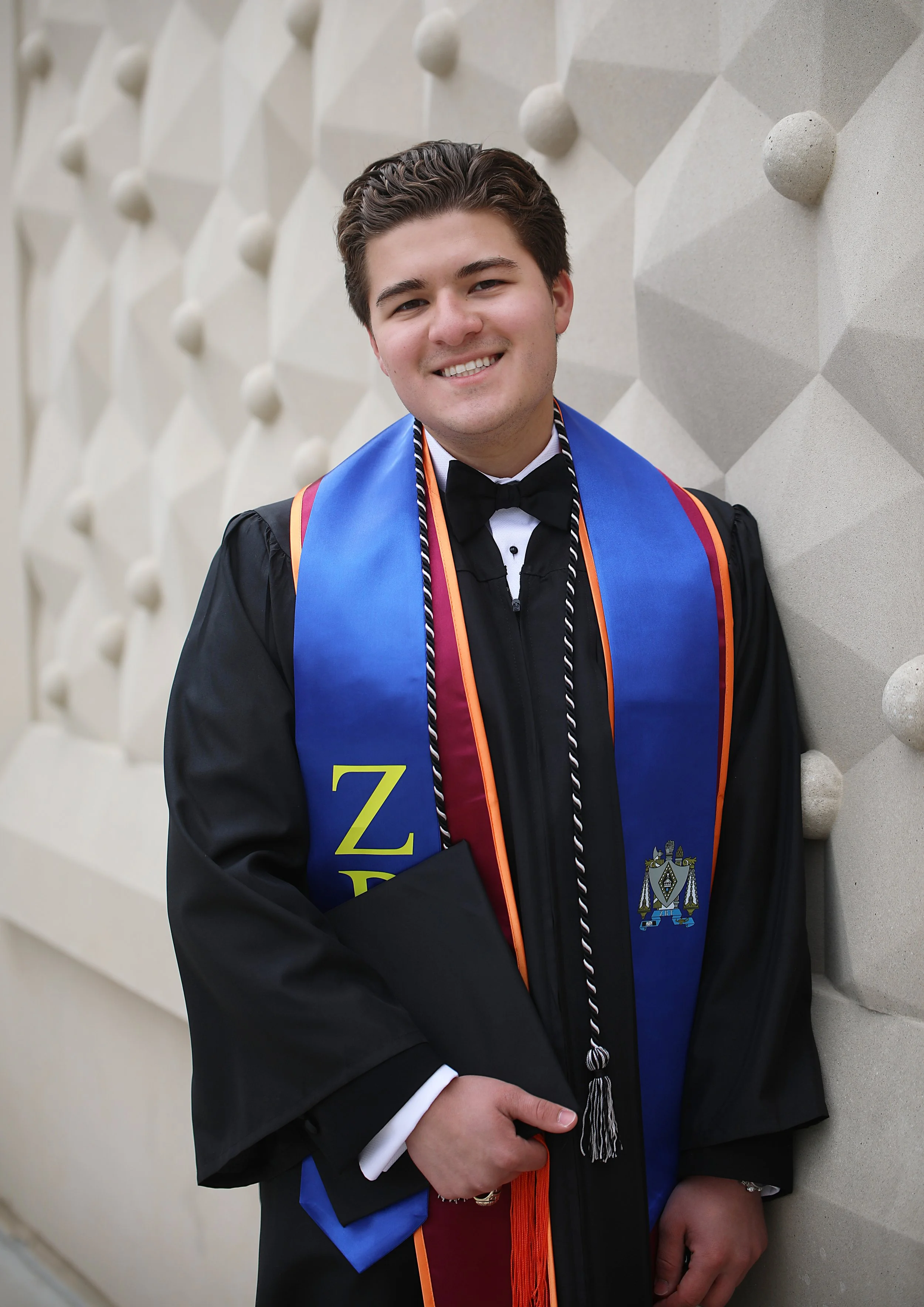 A young man in graduation gown and cap, smiling, wearing honor cords and a regalia stole, standing against an abstract wall with geometric patterns.